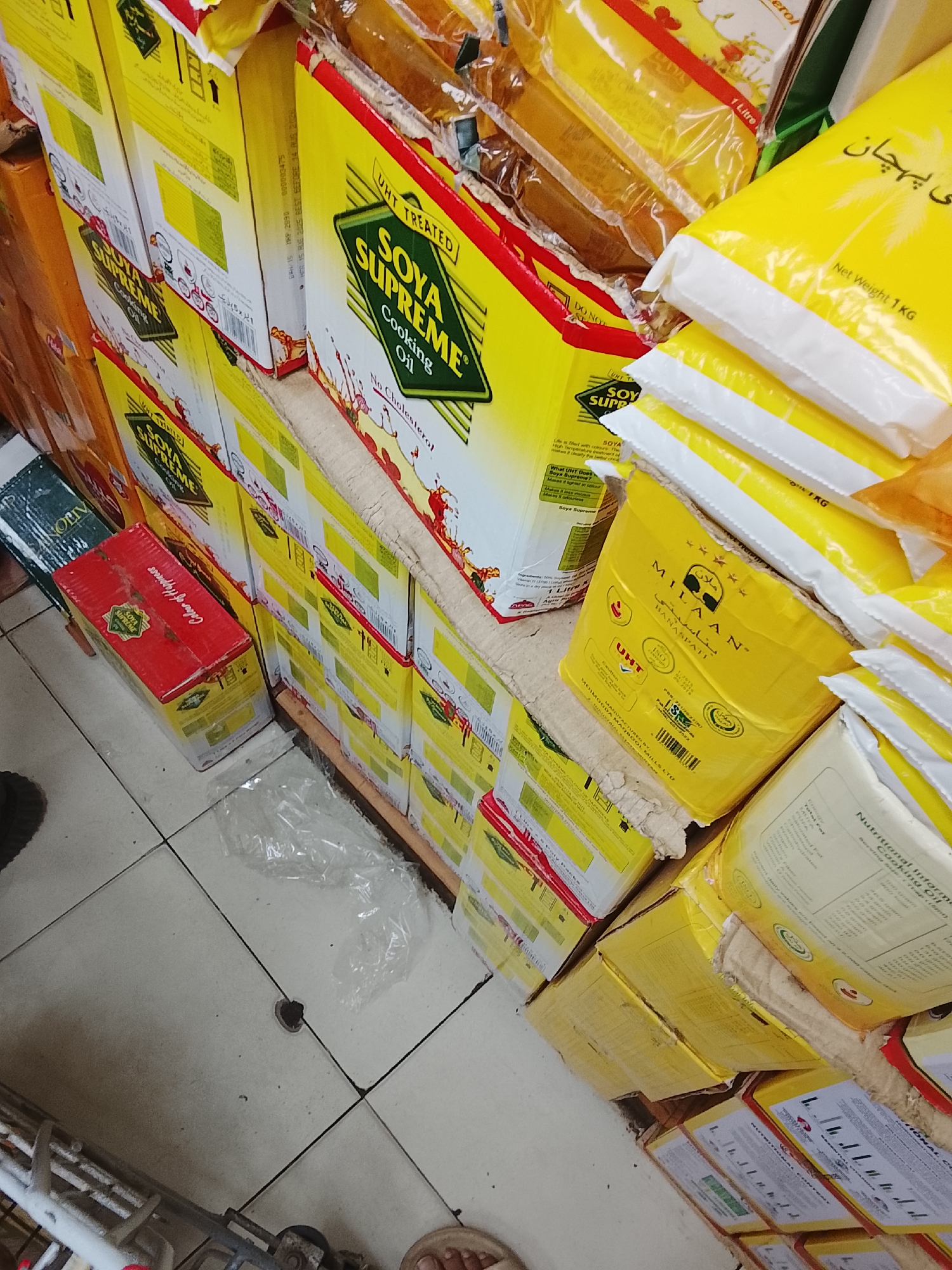 This image, likely taken at ground level, depicts stacks of cooking oil products in what appears to be a retail environment in Gulgasht Colony, Pakistan, based on the presence of Urdu script on the packaging. The dominant brands are "Soya Supreme Cooking Oil" and "MILAAN Banaspati". The oil is packaged in boxes, with some products also in plastic bags. A red box labeled "Cedar of Happiness" is visible on the lower left. The items are arranged on what appears to be a wooden pallet or platform, situated on a tiled floor. A piece of clear plastic packaging material is visible on the floor. Parts of a metal trolley or cart can be seen in the lower left corner, and a person's foot is visible in the lower right corner.