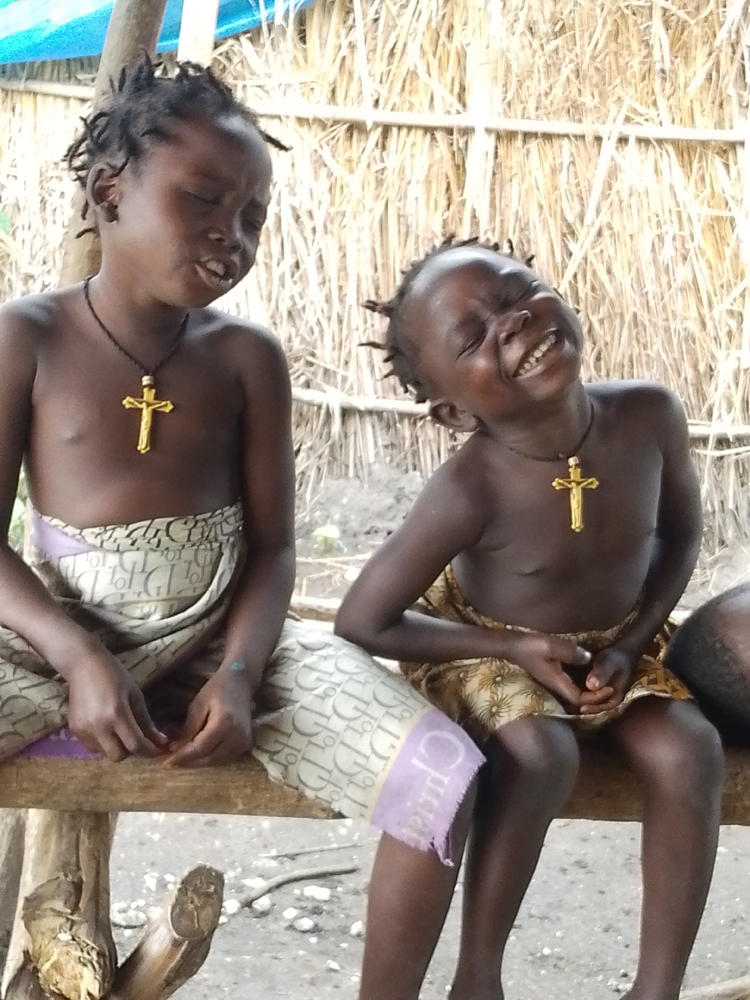 Two young children, likely siblings, are sitting on a wooden beam in front of a thatched wall. The child on the left is looking down and to the left, with eyes closed and mouth slightly open. They are wearing a dark necklace with a golden cross pendant and a patterned skirt. The child on the right is laughing, head tilted back, with eyes closed and mouth wide open, exposing their teeth. They are also wearing a dark necklace with a golden cross pendant and a patterned skirt. The background is a woven wall made of dry grass or reeds. The ground beneath them appears to be dirt with small stones. This scene is located in Otulu, Nigeria.