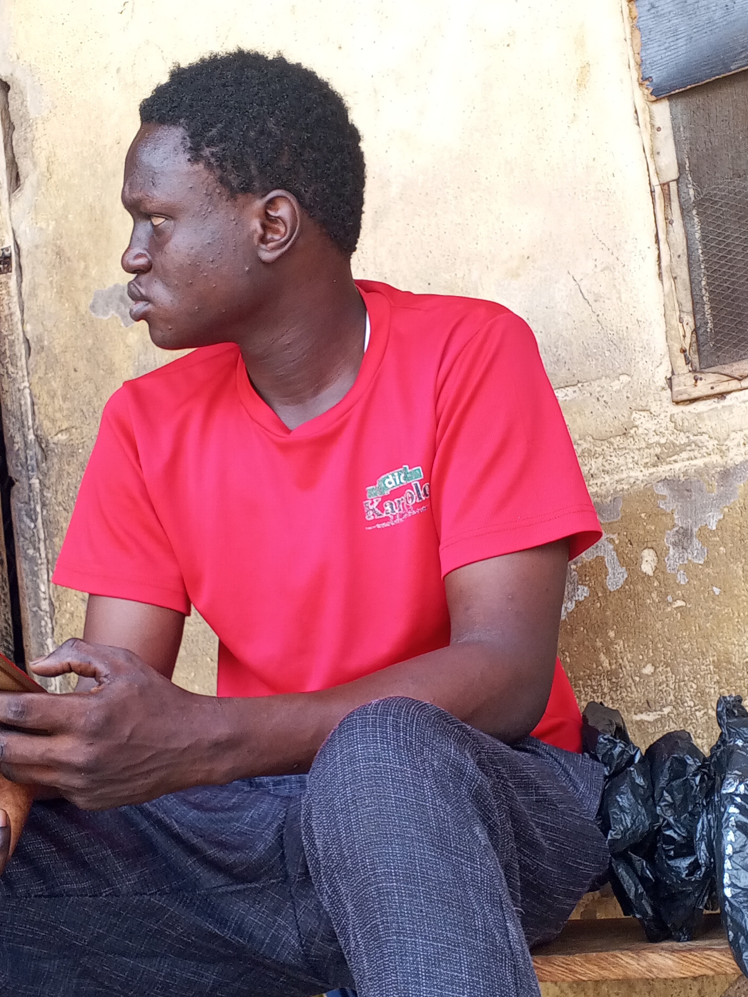A medium shot depicts a dark-skinned man seated against a weathered, light-colored wall. He is wearing a bright red t-shirt with a logo visible on the upper left chest, and dark, patterned pants. His arms are crossed, and he holds a phone in one hand. His gaze is directed to the left. A black plastic bag sits near him on what appears to be a wooden surface. A partially visible window with what appears to be a mesh screen is in the upper right. The location appears to be an outdoor, possibly urban setting, in the city of Unknown, Unknown.