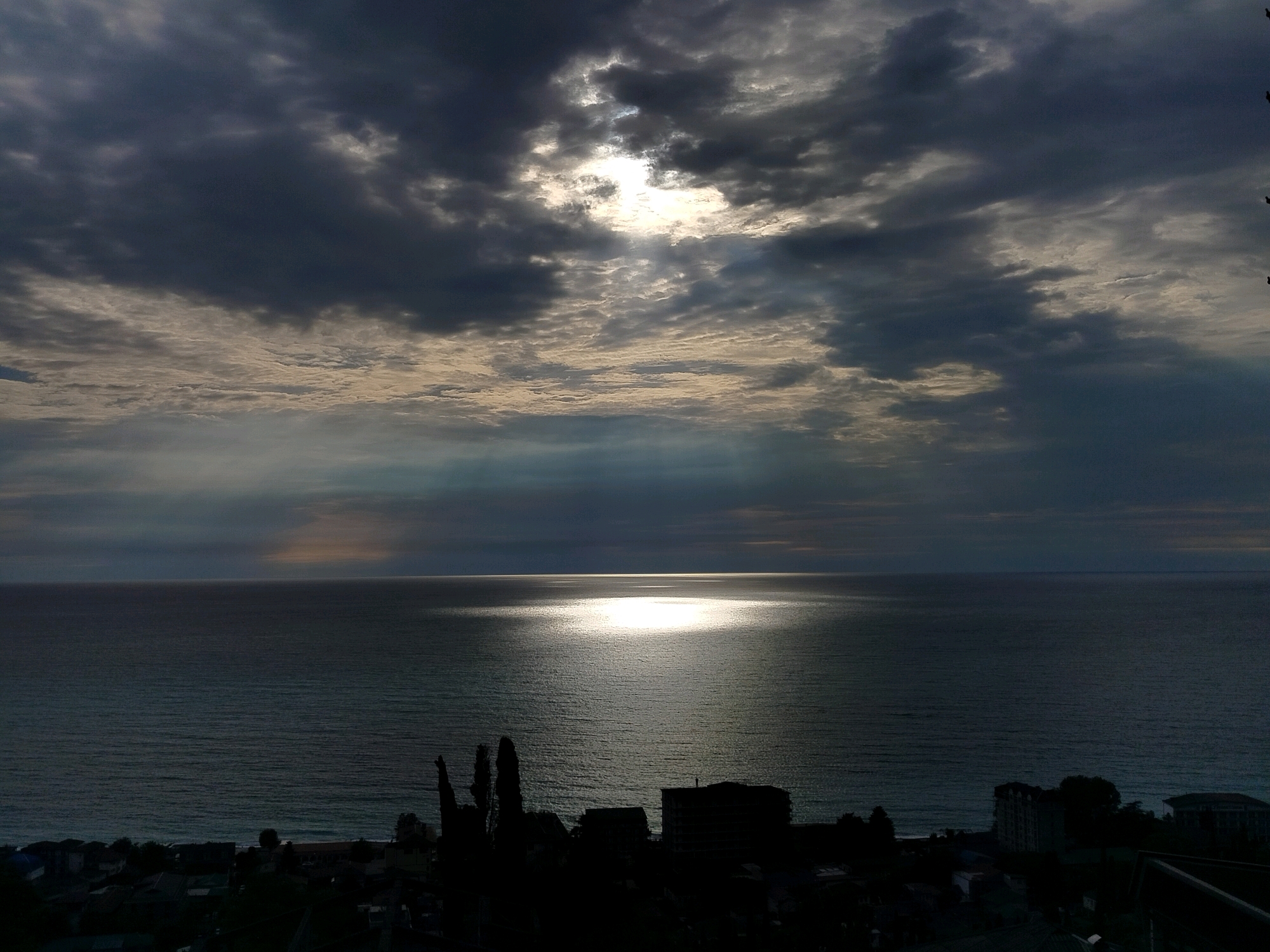 An aerial, wide-angle, high-definition, low-light photograph captures the sea and a darkening sky over a silhouetted cityscape in Gagra, Abkhazia. Dark, heavy clouds dominate the sky, but a break reveals the sun, its rays piercing through, casting a shimmering, bright reflection on the surface of the water. The sea appears calm with gentle ripples. In the foreground, the dark silhouettes of trees and buildings are visible along the coastline, suggesting a dusk or pre-storm atmosphere. The overall scene is dramatic and moody, with a stark contrast between the light on the water and the encroaching darkness of the sky and land.