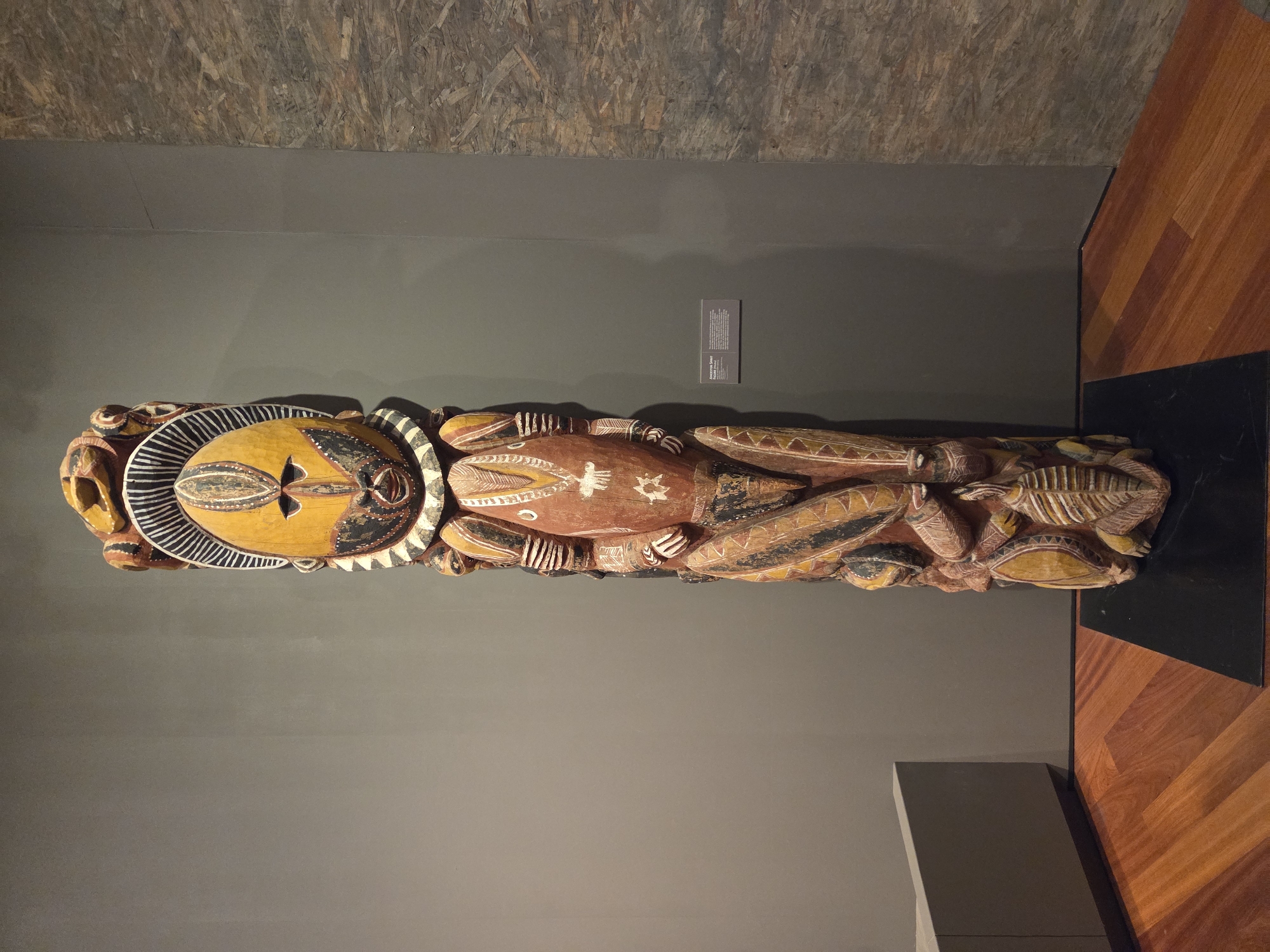 The image shows a large, intricately carved wooden sculpture of a stylized human figure, standing upright on a black rectangular base. The sculpture is positioned against a plain grey wall with a textured panel to its right. The figure is adorned with vibrant colors, primarily yellow, red, black, and white, in geometric patterns and depictions of human features, such as a large face with distinct eyes and a mouth. The lower portion of the sculpture is adorned with multiple smaller carved elements that appear to represent limbs or other symbolic forms.
To the left of the sculpture is a low, grey, angular object, possibly a display case or seating. The floor is made of polished wooden planks with a warm, reddish-brown hue, laid in a diagonal pattern. The overall setting suggests an indoor exhibition space, likely a museum or gallery.
A small informational plaque is visible on the grey wall to the right of the sculpture, indicating this is an "Ancestor Spirit" sculpture. This suggests a cultural or historical context for the artwork. The lighting appears to be artificial and evenly distributed, highlighting the details of the sculpture. The composition is centered on the sculpture, with the background elements providing context but not distracting from the main subject.
This scene is located within the city of Santa Ana, United States of America.