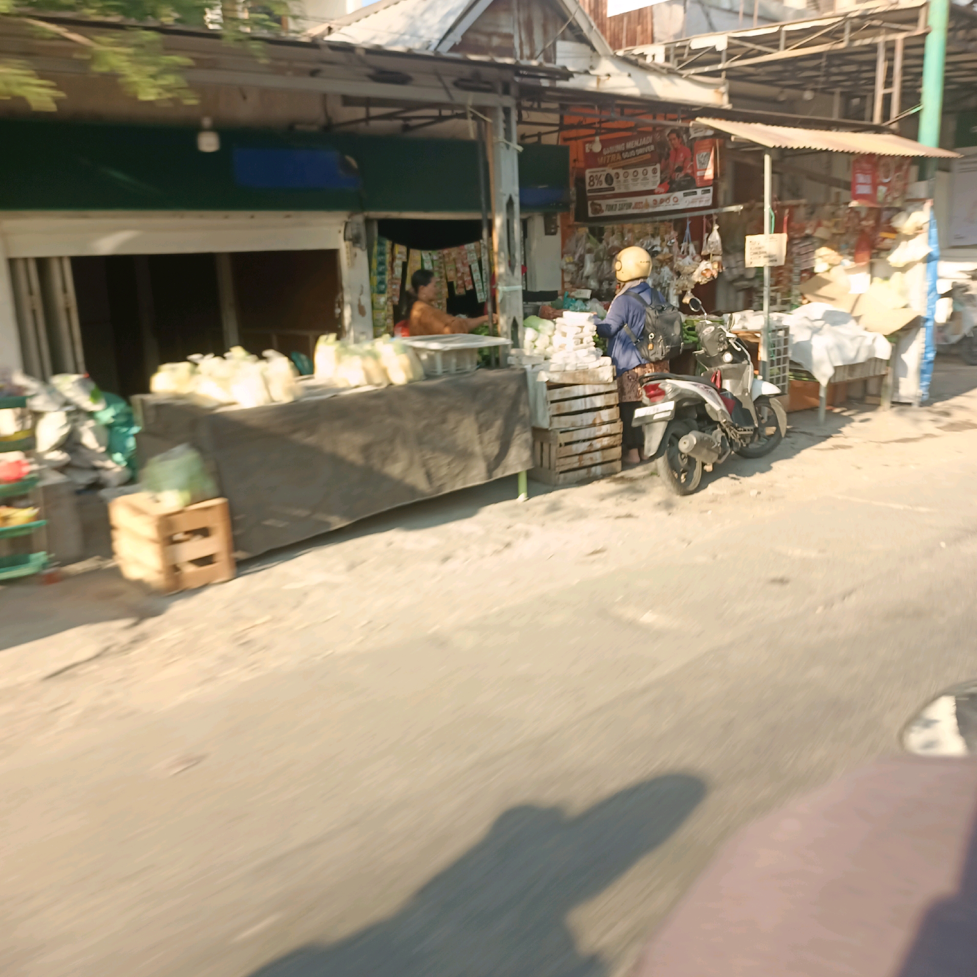 The image depicts a street scene in Surabaya, Indonesia, specifically in front of a small shop or market stall. A person wearing a blue jacket and a yellow helmet is on a motorcycle. The motorcycle is parked next to a display of stacked wooden crates, which hold what appear to be white plastic containers. Behind the crates, a stall with a dark tarp is laden with white sacks, likely agricultural produce. To the left of the stall, another person in an orange shirt is visible inside a small building, possibly the shop owner. The background shows the typical architecture of a street-side establishment in Indonesia, with a corrugated metal roof and various advertisements. The ground is paved with asphalt. The overall impression is of a busy market area on a sunny day.