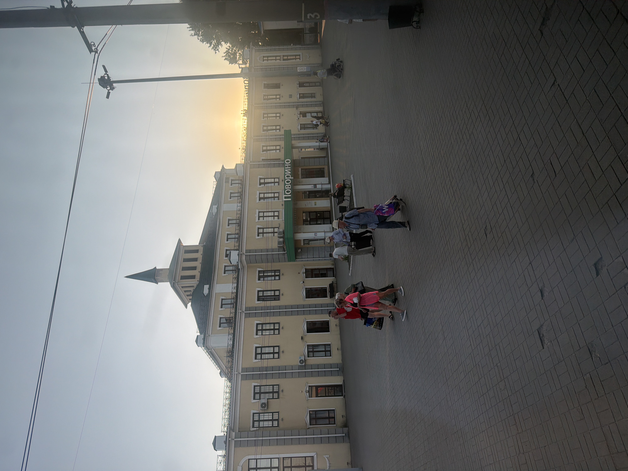 The image depicts the exterior of a light yellow, two-story building that appears to be a train station, with a prominent green sign bearing the Cyrillic text "ПОВОРиНО" (POVORINO). The building has a central section topped with a dark green, spire-like structure. Several windows with dark frames are visible on both levels, along with air conditioning units.

In front of the building, a paved plaza is occupied by several individuals. From left to right: a couple walks towards the viewer, the woman in a bright pink dress and the man in a red shirt and dark shorts, both carrying bags. Further right, a group of people are walking, including a person in a wheelchair. To the far right, a tall street light pole with multiple fixtures is visible, along with a dark trash bin. The sky is a pale blue, with the sun setting on the horizon, casting a warm glow. The overall scene suggests a train station in the late afternoon or early evening. The location is identified as the city of Krasnoe, Russia.