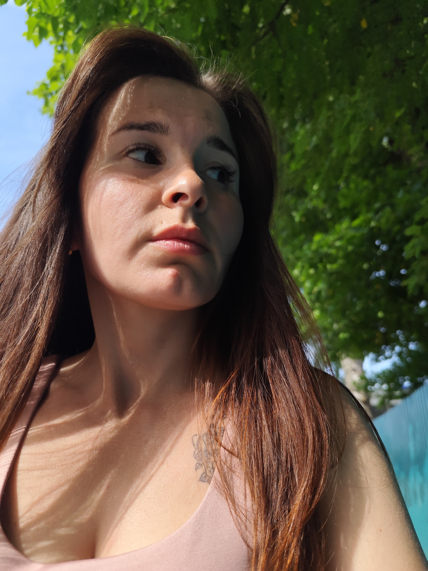 A woman with long, brown hair is pictured outdoors. The photo appears to have been taken in a location with trees, as green foliage is visible in the background, with a glimpse of blue sky. The woman is wearing a light pink top and is gazing to the left. The lighting suggests a sunny day, with shadows cast across her face and chest from the trees overhead. A small tattoo is visible on her upper chest. It is not possible to determine the exact location as Фоки, Russia, based on the image.