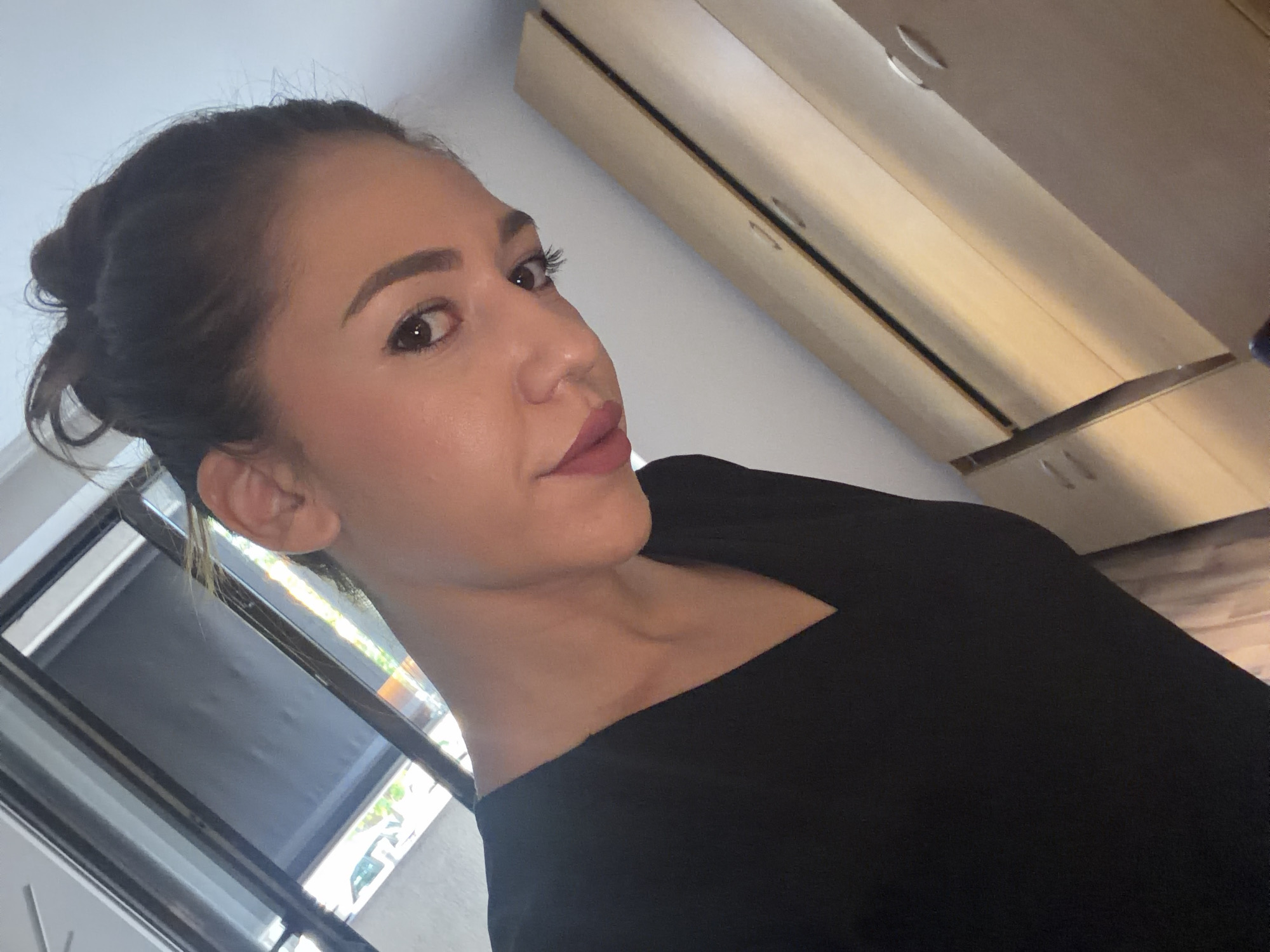 A woman is captured in a selfie-style photo indoors. She is wearing a black V-neck shirt. Her dark hair is pulled up in a bun. The background includes a window with blinds, suggesting an interior setting. The lighting suggests it is daytime. The image does not provide information about the city of Sofia, Bulgaria.