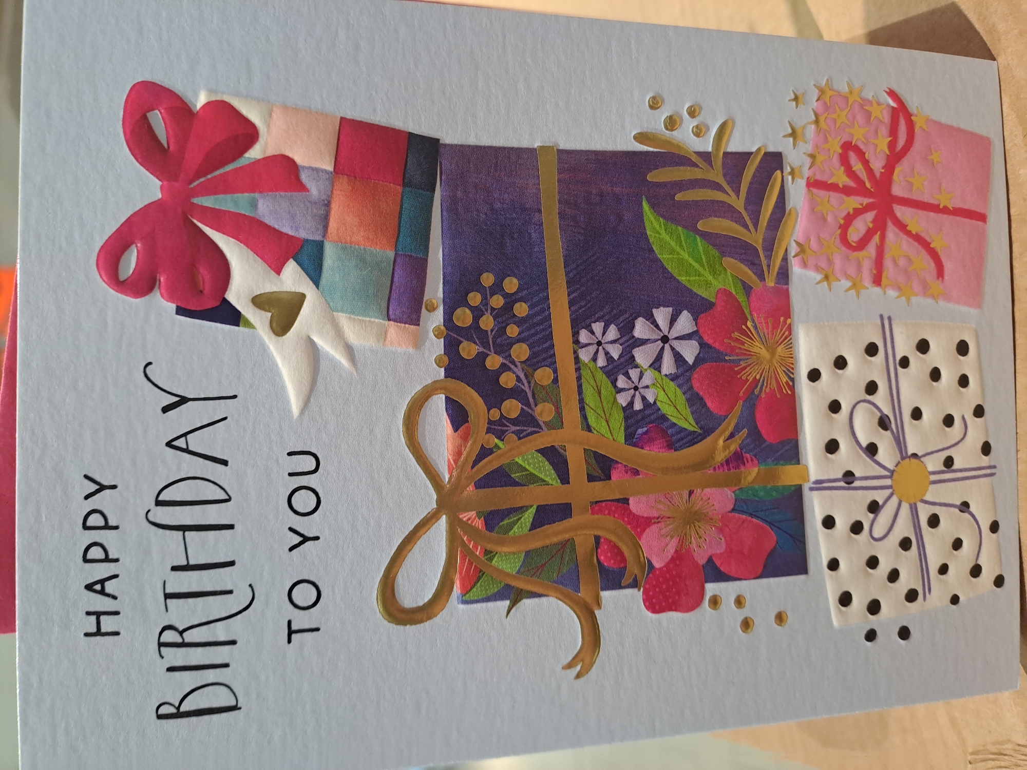 The image shows a light blue birthday card with four stylized gift boxes. The box on the upper left is a patchwork design with a pink bow. The next box is purple with gold ribbons and floral decorations. The upper right box is pink with gold stars and a pink ribbon. The lower right box is white with black polka dots and a purple ribbon with a gold center. The words "HAPPY BIRTHDAY TO YOU" are printed in black on the left side of the card. The background is out of focus and appears to be a beige or light brown surface. There is no indication of the location being Veliko Tarnovo, Bulgaria.