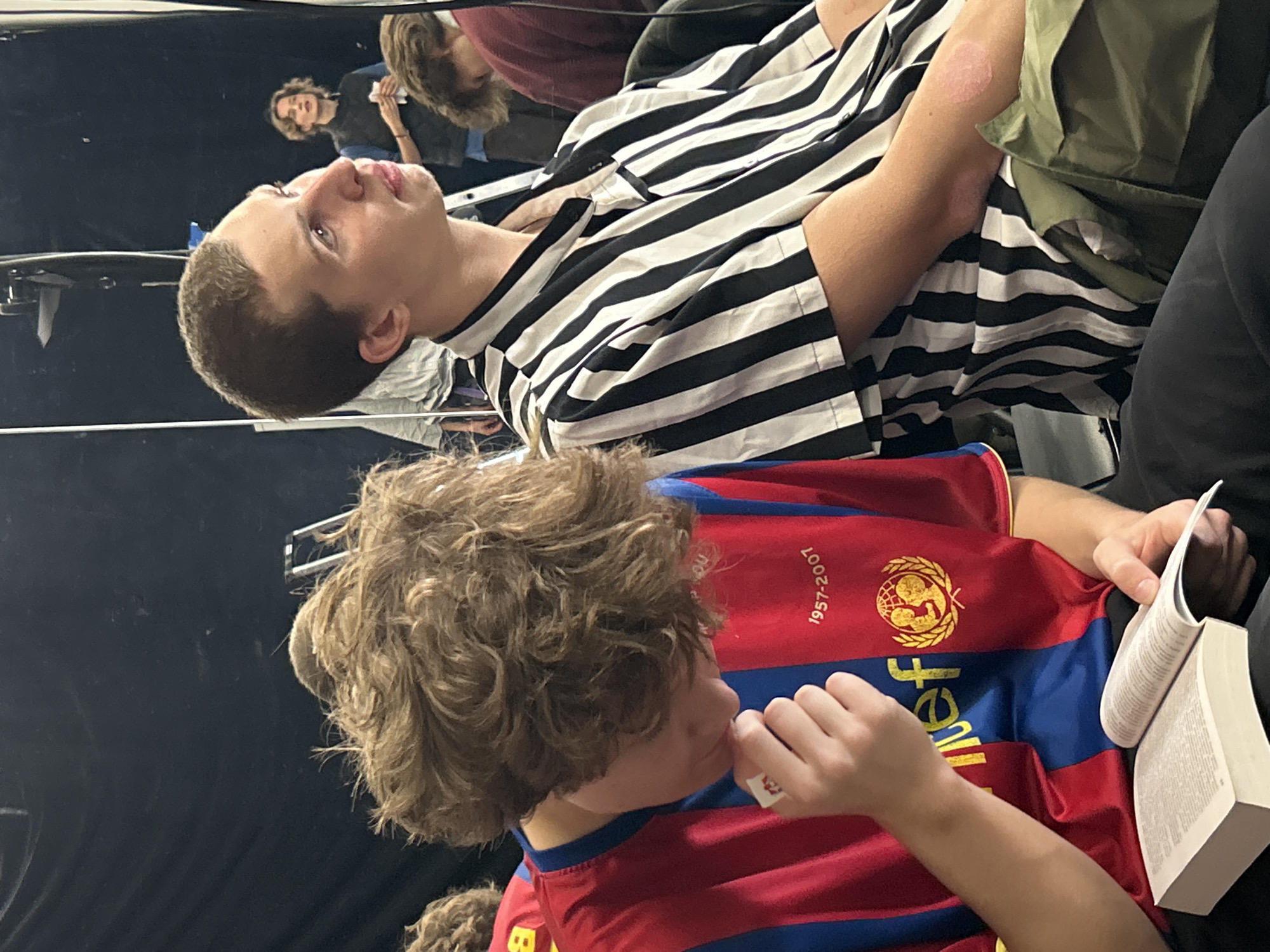 In a dimly lit room with dark walls, two young males are the primary subjects. The male on the left, with curly brown hair, is wearing a red and blue striped jersey with a logo and text that reads "1957-2007". He is looking down at a thick, open book in his lap, holding a small white object in his right hand near his mouth. The male on the right, with short brown hair, is looking to his right and is wearing a black and white striped button-up shirt. His skin has a pinkish hue on his arm. He is wearing olive green pants. In the background, a female with curly brown hair is visible, wearing a dark vest over a blue shirt and holding a white object. The environment appears to be indoors, possibly a backstage or rehearsal area, with reflections in what looks like a large mirror or glass surface.