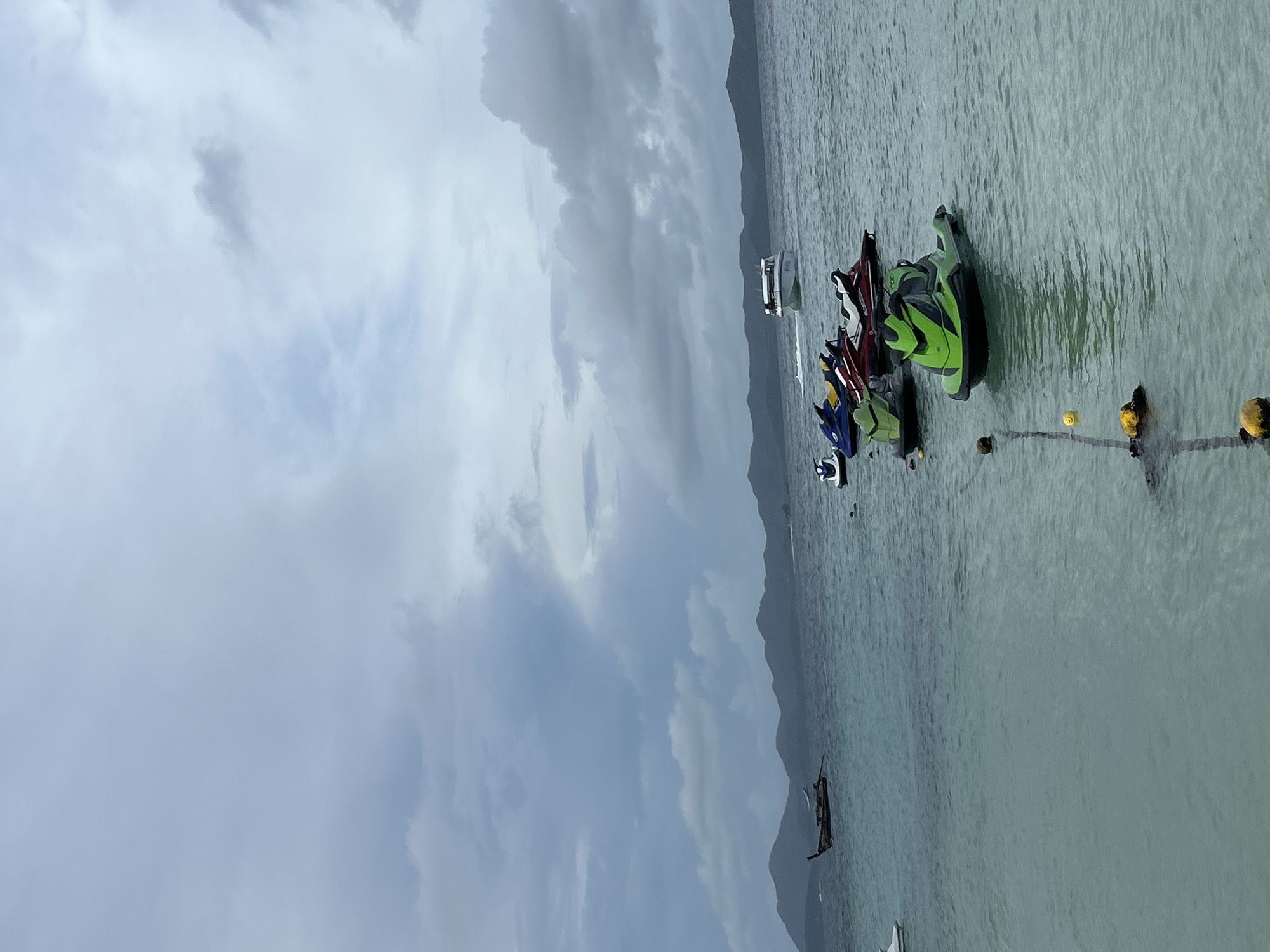 The image depicts a tranquil scene of the ocean near Tambon Phru Nai, Thailand. The water is a clear turquoise, with visible buoys and a rope marking a boundary. Several jet skis are moored in the water, with three in the foreground and several more further back. In the distance, a longtail boat navigates the water, with a speedboat approaching from the right. The horizon is lined with lush green hills, shrouded in a light mist under a cloudy sky. The overall atmosphere is serene and peaceful, conveying a sense of a quiet day at sea.