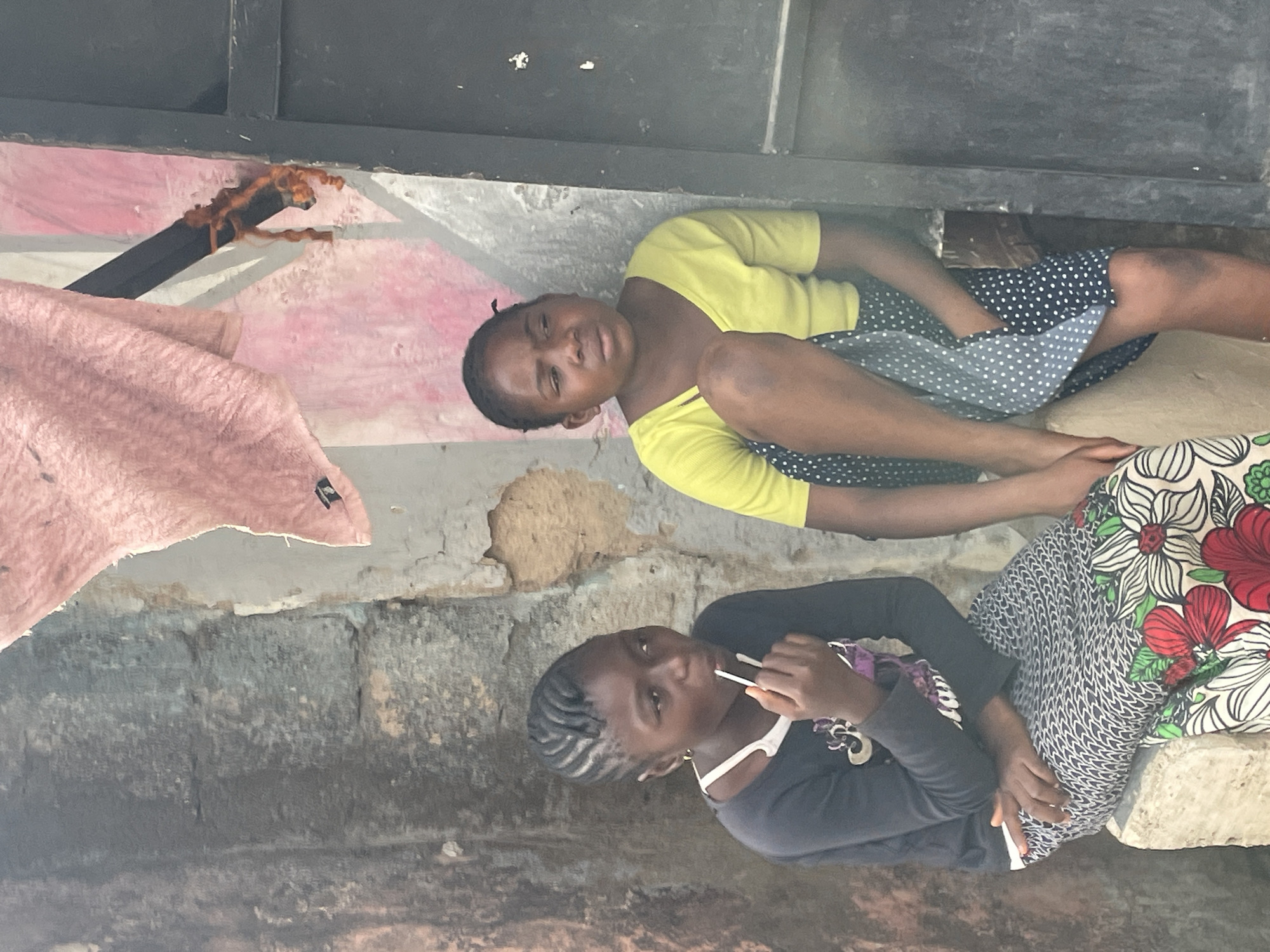 This image, set in Jos, Nigeria, features two young girls seated side-by-side on a low concrete ledge or step, looking towards the viewer. The girl on the left wears a bright yellow short-sleeved top and a dark blue skirt with white polka dots. She has dark hair and a calm expression. Her legs are bent, and she appears to be resting comfortably.
Beside her, on the right, sits another girl dressed in a dark long-sleeved top and a skirt featuring a vibrant floral and geometric pattern in red, green, white, and black. She has braided hair (cornrows) and is holding a white stick-like object, possibly a lollipop or candy, to her mouth. Her expression is also neutral as she looks towards the camera.
The background consists of a worn wall, likely concrete or brick, with sections of peeling light-colored paint or plaster, revealing the rough texture beneath. Above the girls, a darker structure, possibly a metal beam or frame, extends horizontally. A black pipe or bar is visible to the upper left, partially covered with reddish-brown fibrous material. A light pink fabric or blanket is draped over part of the wall on the far left. The natural lighting indicates it is daytime. There is no visible text in the image.