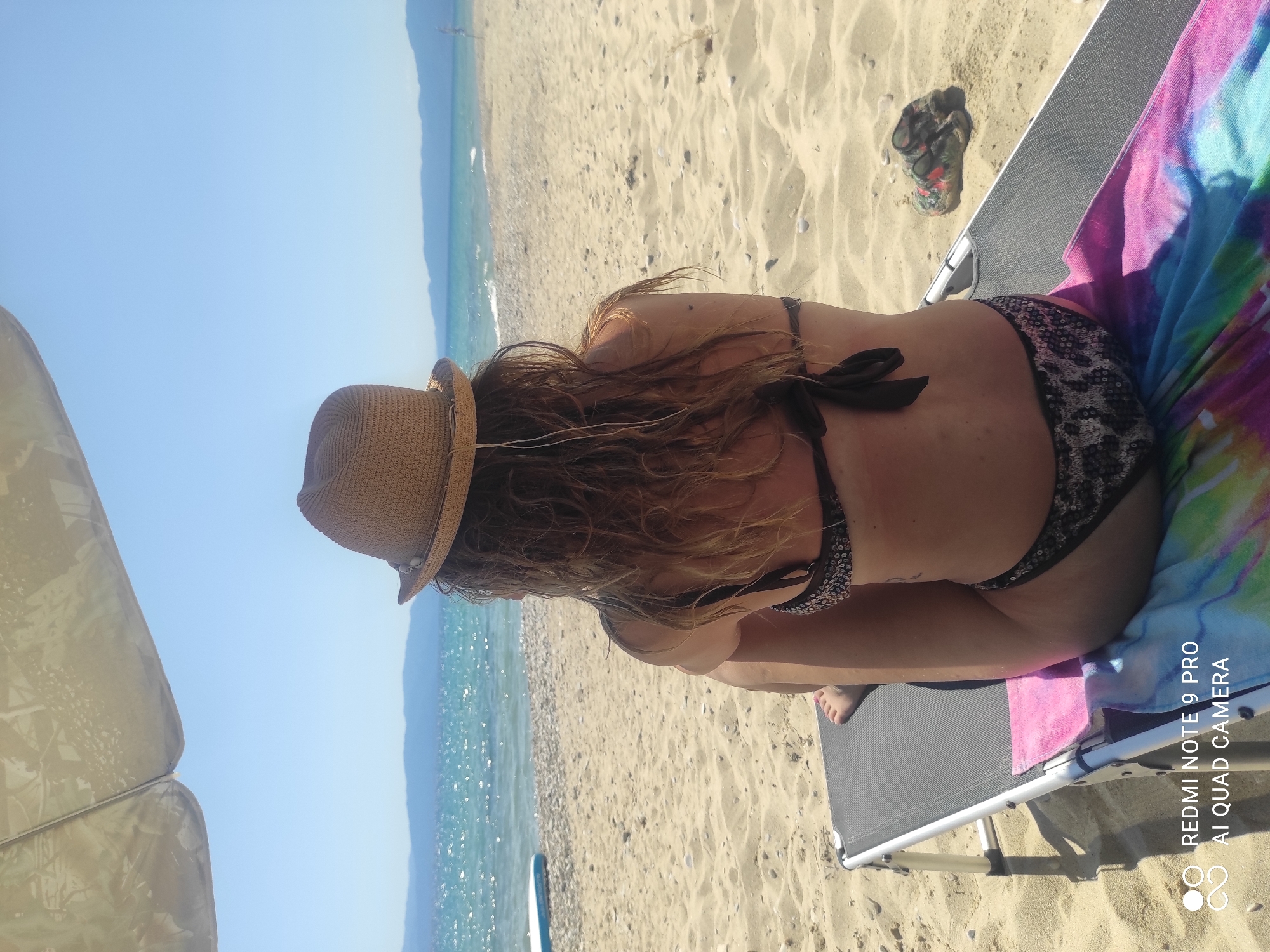 A woman wearing a straw hat and a patterned bikini sits on a beach chair on Ofrynio Beach, Greece. She is facing away from the camera, looking towards the ocean. The beach is sandy, with pebbles and shells visible along the waterline. The water is calm and blue, with distant mountains on the horizon. The sky is clear and sunny. A beach umbrella provides shade over the woman. There are sandals lying on the sand to the right of the chair. The image also includes a watermark at the bottom left corner indicating "REDMI NOTE 9 PRO AI QUAD CAMERA".