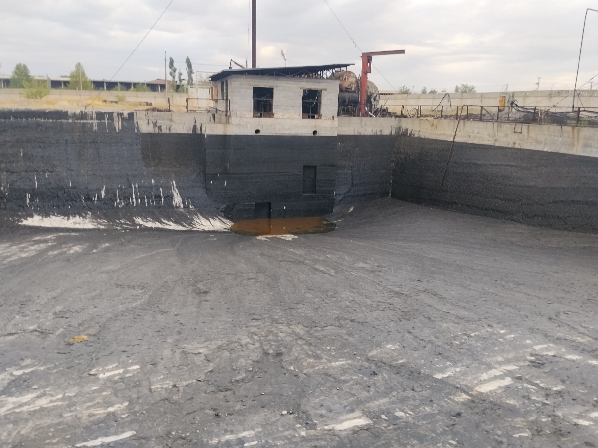 The image shows a large, open, concrete basin with dark, sludgy material lining its bottom and sides. The basin appears to be a settling pond or a similar industrial water treatment facility. In the background, there are industrial buildings and structures, along with some trees and a cloudy sky. A small, dilapidated building with open window frames is situated on the edge of the basin. The ground in the foreground is a mixture of dirt and debris, showing tire tracks. The scene suggests an industrial area that may be under construction, disused, or undergoing maintenance. The location is identified as Manas, Kyrgyzstan.
