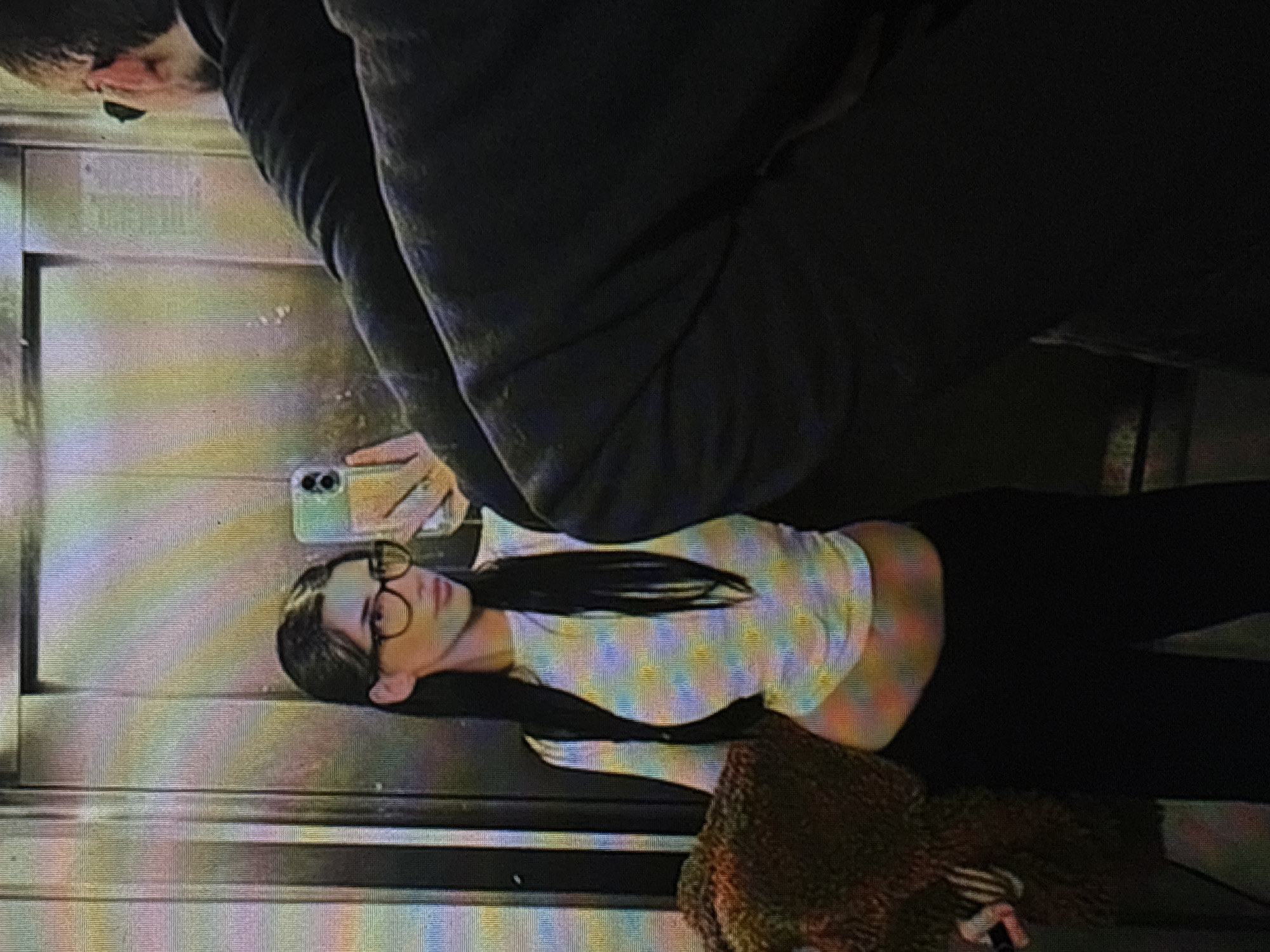 A selfie taken in what appears to be an elevator. A person with long dark hair, wearing glasses, a crop top, and dark pants is visible. Another person is partially visible in the upper portion of the frame, wearing a dark-colored garment. A brown, textured object is in the lower right corner, possibly a purse. The elevator interior appears to be metal. There is no contextual information available to determine the location in Sofia, Bulgaria.