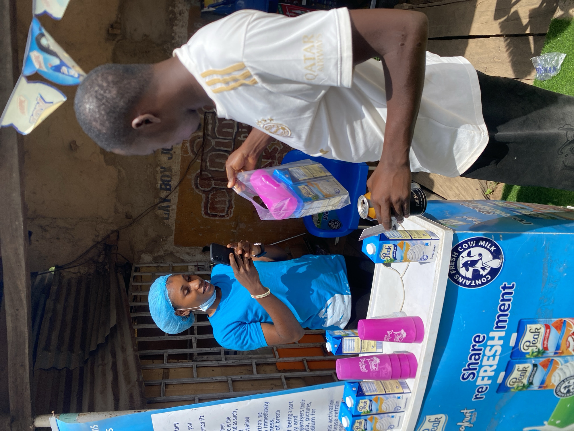 In Bukuru, Nigeria, a woman wearing a blue hairnet and t-shirt is standing behind a blue refrigerator displaying "Peak" yogurt drinks. She is looking at her smartphone. A man in a white t-shirt with gold stripes and "Qatar Airways" printed on it is holding a plastic bag containing two yogurt drink cartons and a pink cup. He is standing in front of the refrigerator, interacting with the woman. The scene appears to be an outdoor stall with a corrugated metal roof overhead. A banner with text is visible on the left side of the frame.