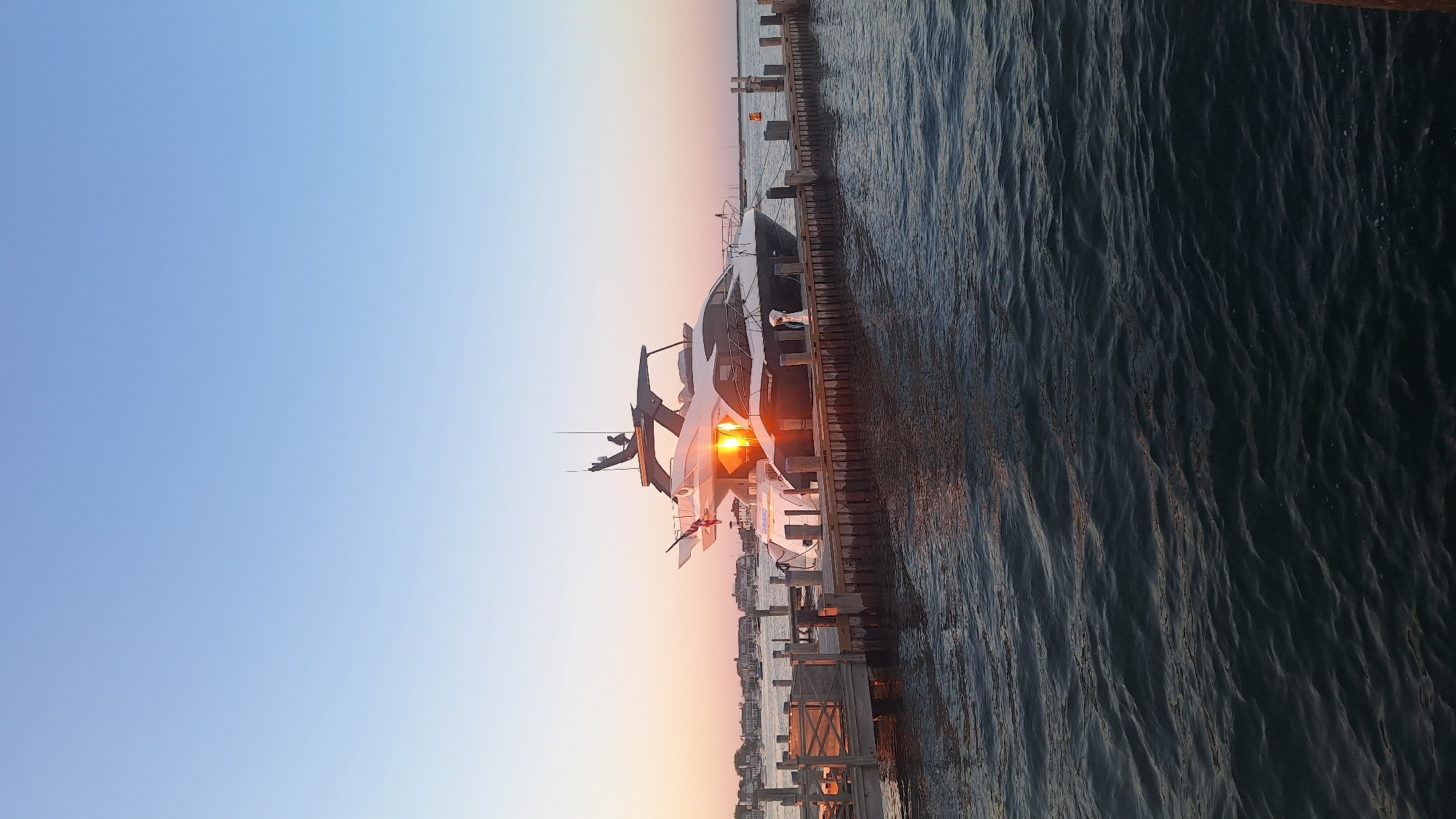 A large white and black yacht is docked at a wooden pier. The yacht has multiple levels and a dark roof. The sun is setting, casting an orange glow on the yacht and the water. The sky is a gradient of blue and pink. In the background, there are houses and a few other boats. The water is calm with ripples. This scene is likely in Nantucket, Massachusetts, a popular destination for yachts and coastal living.