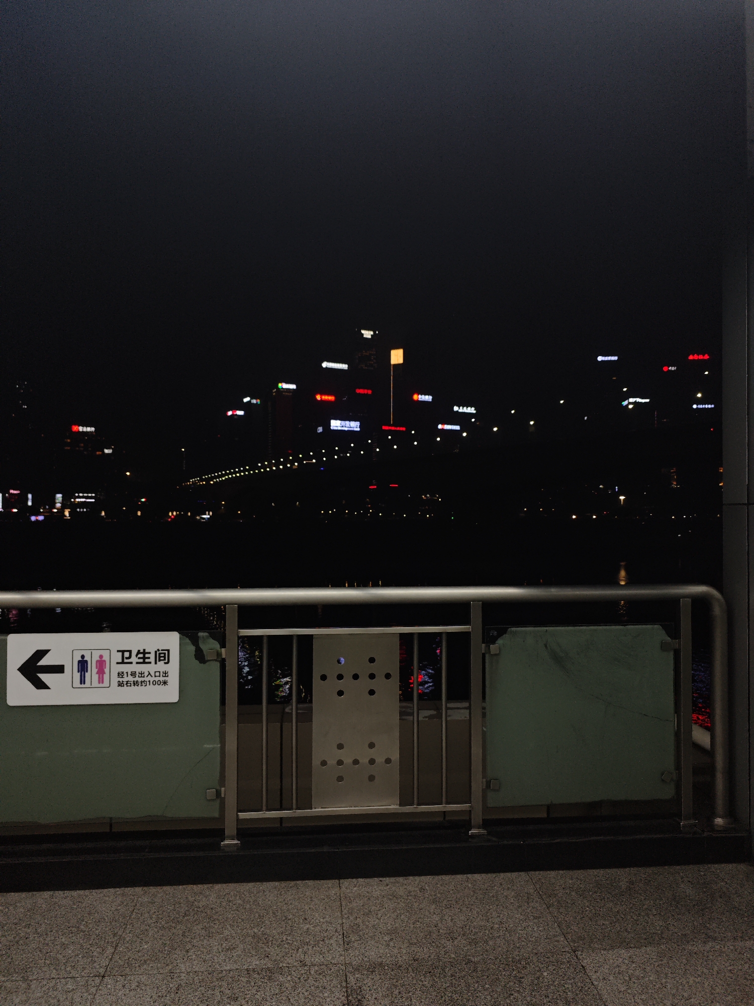 A nighttime urban scene. A railing separates the foreground from a dark body of water with city lights reflected in it. Behind the water, city buildings are visible, lit up with signs in Chinese. A sign on the railing points to a restroom. The location is Yuzhong District, China.