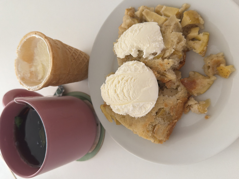 A high-angle photograph captures a dessert arrangement on a white, unadorned surface. In the upper left, a pink ceramic mug contains a dark liquid, possibly tea, with several small dark green leaf fragments visible on its surface. This mug rests on a circular coaster, which is predominantly green with a lighter edge. To the right of the mug, a light brown, textured waffle cone stands upright, containing a partially melted, pale yellow substance, consistent with ice cream. The upper rim of the cone shows an irregular, broken edge. In the foreground, centered on a plain white round plate, is a portion of a baked dessert, identifiable as a fruit crumble or pie, likely apple, given the visible chunks of light yellow fruit within a golden-brown baked crust. Two scoops of white ice cream top the dessert; the scoop on the left is largely intact, while the scoop on the right appears partially melted or fragmented. The environment is devoid of individuals or other discernible objects beyond the described items. The specific geographical location within Tel-Aviv, Israel, cannot be determined from the visual information presented.