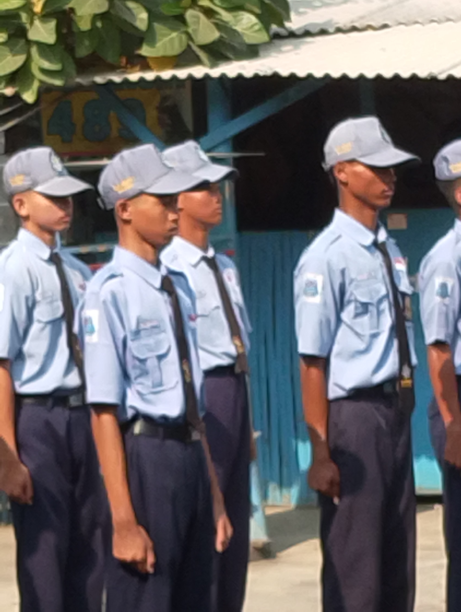 A group of four young males, appearing to be students, stand in formation outdoors. They are all wearing light blue collared shirts, dark blue pants, and grey caps with a logo on the front. Each individual also wears a black tie. The individuals are positioned side-by-side, facing forward and to the right of the frame. In the background, there is a white overhang with a scalloped edge and a blue structure with the number "489" visible, painted in yellow. The ground appears to be paved. This scene is located in Manaruwi, Indonesia.