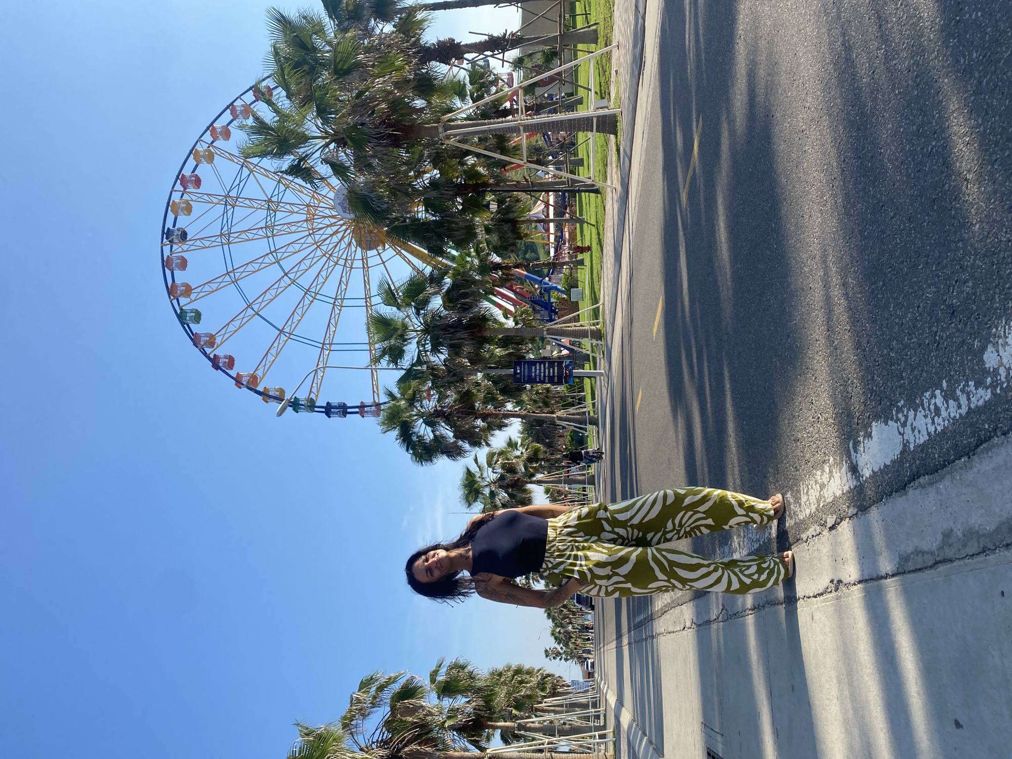 The image captures a full-body, outdoor shot of a woman standing on a paved walkway next to a road. She is facing towards the camera with her hands in the pockets of her green and white patterned pants. She is wearing a dark blue sleeveless top. Behind her, a vibrant Ferris wheel with multi-colored cabins stands tall against a clear blue sky. A row of palm trees lines the walkway and the road, with some of them appearing to be supported by white scaffolding. The road has yellow lane markings and is partially shaded by the trees. In the background, other amusement park structures and a few vehicles are visible, suggesting a lively recreational area. The scene appears to be set in Tien Thanh Ward, Vietnam, indicated by the presence of Vietnamese signage on a blue placard to the right of the frame. The overall atmosphere is bright and sunny, suggesting daytime.