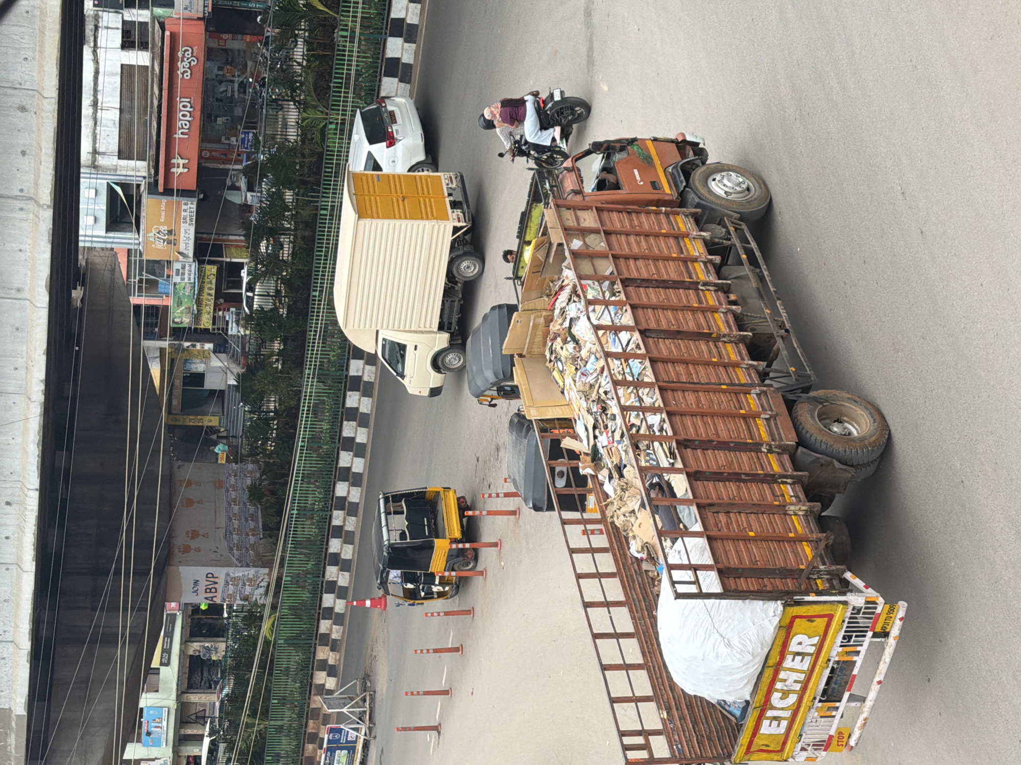 The image captures a street scene in Shamshabad, India, under an elevated overpass. In the foreground, a large, rusted brown truck filled with garbage is parked or moving slowly. The truck's rear displays the "EICHER" brand and an "AP31TU 9509" license plate, along with a "STOP" sign. Beside the truck, a large white tarp-covered object is visible. Behind the truck, a yellow and black auto-rickshaw is stopped, and a black auto-rickshaw is also present. Further back, a white panel van and a white SUV are on the road. On the right side of the frame, two individuals on a motorcycle are riding away from the viewer. In the background, buildings line the street, with various storefronts and signage, including "happi" and "Coca-Cola." A green fence separates the road from the sidewalk area. The overall environment appears to be an urban street with moderate traffic.