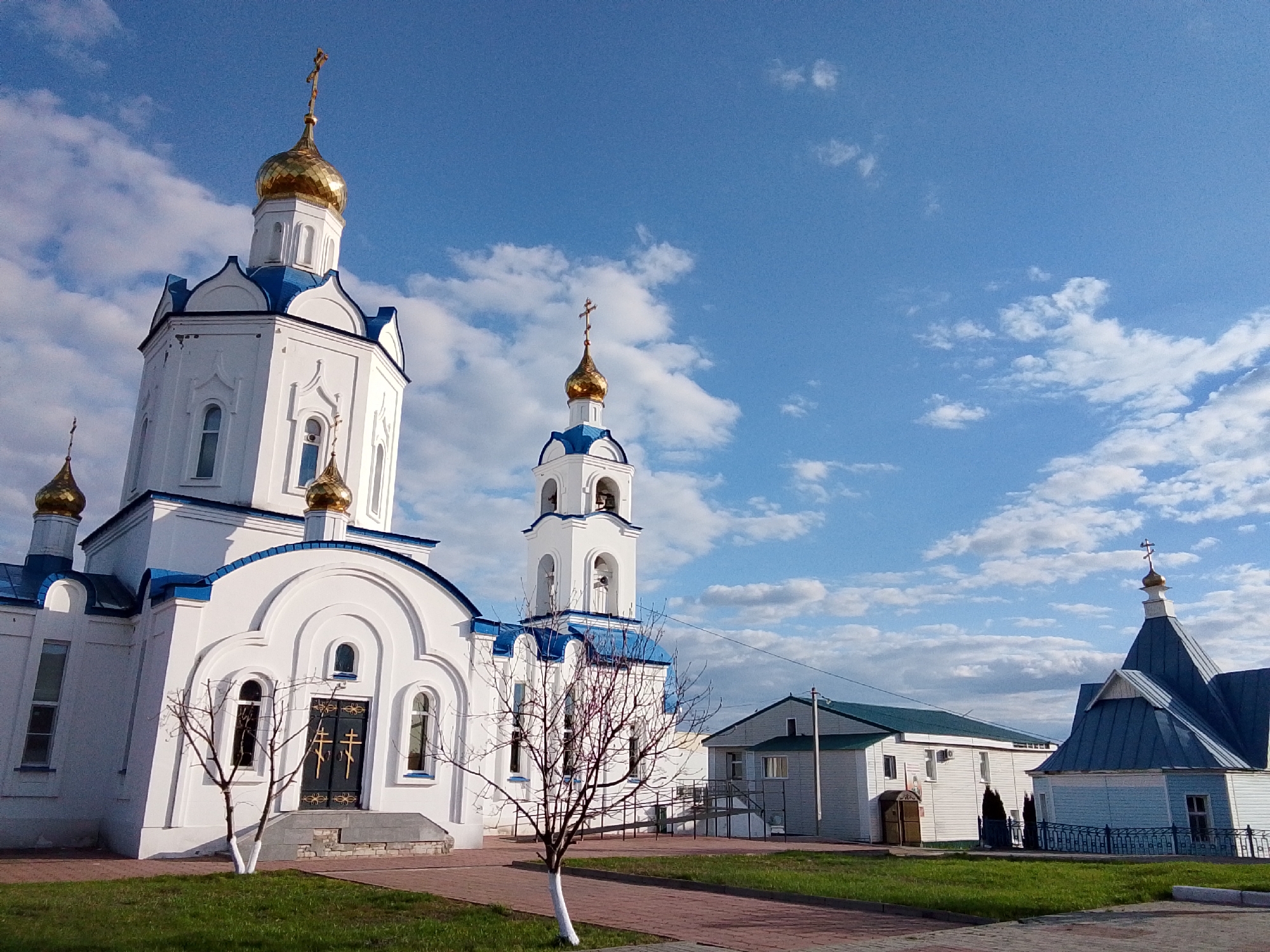 This media file depicts an Orthodox Christian church complex in Khlevnoye, Russia, under a bright blue sky with scattered white clouds. The main subject is a large, white church with blue roofs and multiple golden onion domes, each topped with a cross. A tall, white bell tower, also with blue roofs and golden domes and crosses, stands slightly behind the main church, with bells visible in its open arches.
The entrance to the main church features a dark door adorned with two stylized golden crosses. Paved red-brick pathways traverse a well-maintained green lawn. Several bare trees with white-painted lower trunks are planted around the complex. To the right of the main church, there are two smaller buildings: one is a white structure with a green roof, possibly a rectory or community hall, featuring a small, unreadable sign by its entrance and an outdoor air conditioning unit. The other, further right, is a smaller, light blue wooden chapel with a blue roof and a golden cross.
The scene is brightly lit by natural daylight, indicating daytime. No people are visible in the image. The prominent architectural style, featuring onion domes and crosses, clearly identifies the site as an Orthodox church.