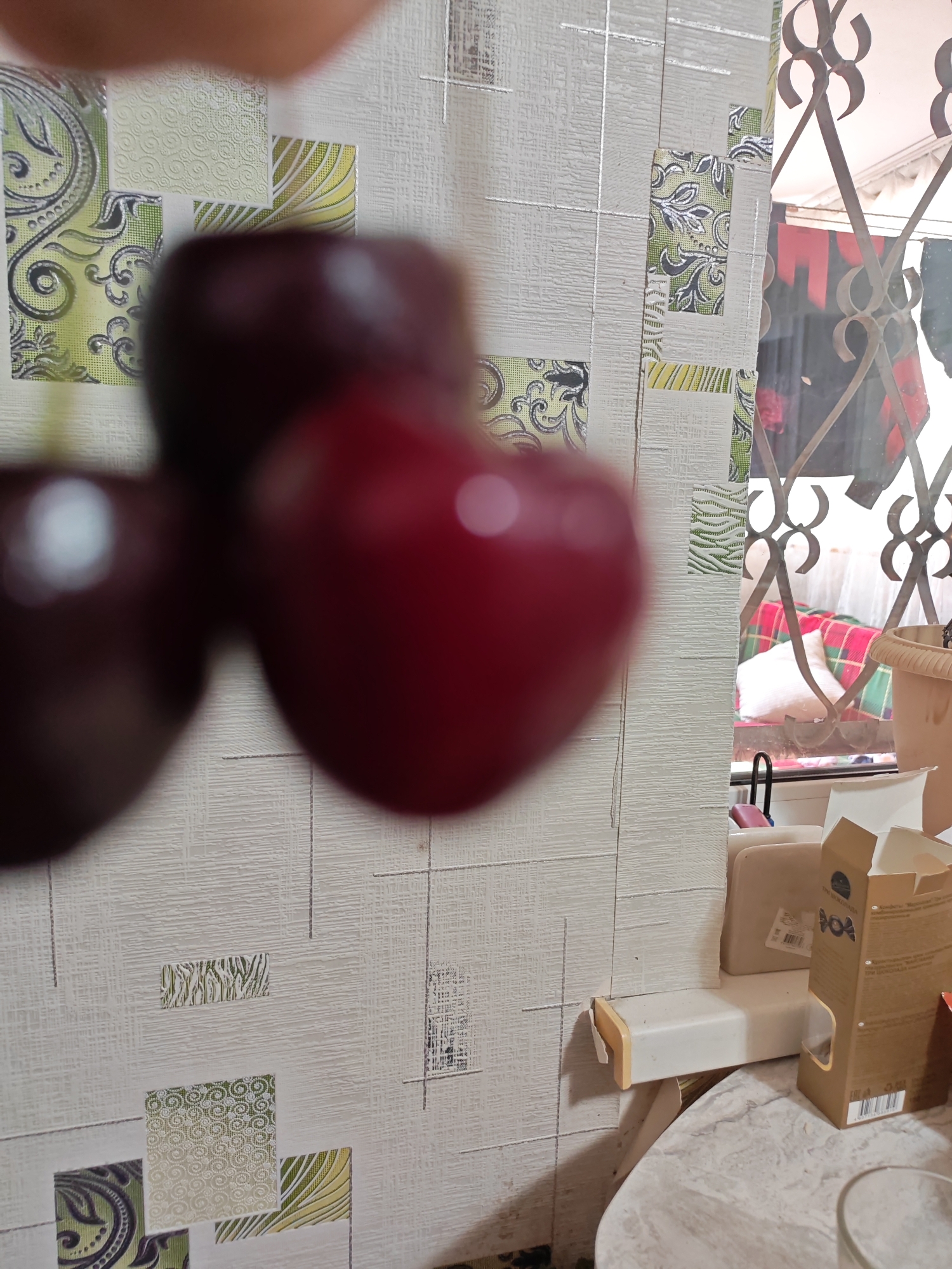 The image shows a close-up of three cherries, out of focus, hanging against a wall with a textured, patterned wallpaper. The wallpaper features a grid of squares, some of which have decorative patterns in green, yellow, and white. To the right of the cherries, a metal grate is visible, suggesting a window or a decorative grille. Through the grate, there are glimpses of laundry hanging and possibly a bed with a checkered blanket. In the foreground on the right, there are some boxes and a beige plastic planter. The scene appears to be indoors, possibly in a kitchen or living area, and the lighting suggests it is daytime. There are no visible people or overt actions. The location is Tolyatti, Russia.
