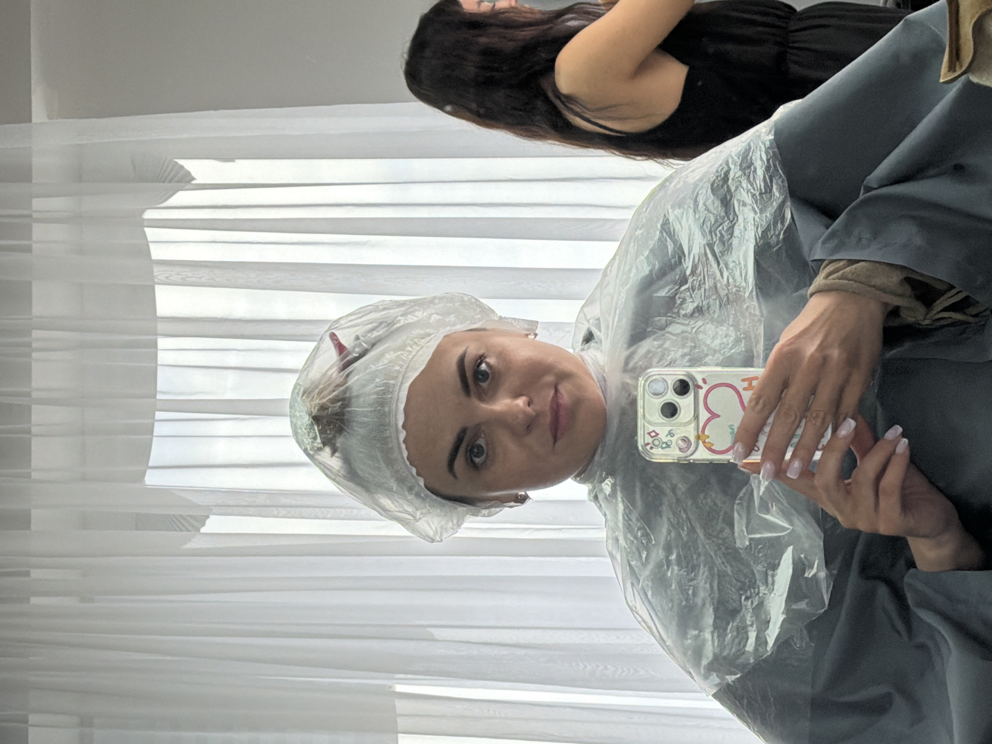 A woman in a clear plastic shower cap and a gray garment is taking a selfie in what appears to be a salon setting. She is holding a smartphone with a decorative case. Another person with long dark hair is visible in the background, attending to the woman in the foreground. White blinds are seen in the background. The setting appears indoors, and the location, Votkinsk, Russia, is provided externally.
