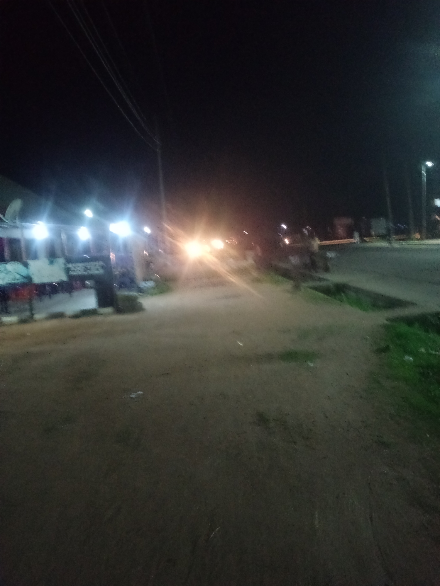 A nighttime street scene in Jalingo, Nigeria, features a dirt road or path in the foreground, leading towards a paved road on the right. Several bright lights illuminate the area from buildings on the left, creating strong glare. Some of these lights appear to be streetlights or lights from shops. The buildings on the left seem to be commercial establishments, with some signs and awnings visible. On the paved road, there are visible headlights of vehicles, suggesting traffic. A few indistinct figures or silhouettes of people can be seen on the right side of the road, near some structures. The overall impression is of a moderately busy urban street at night, with artificial lighting the primary source of illumination.