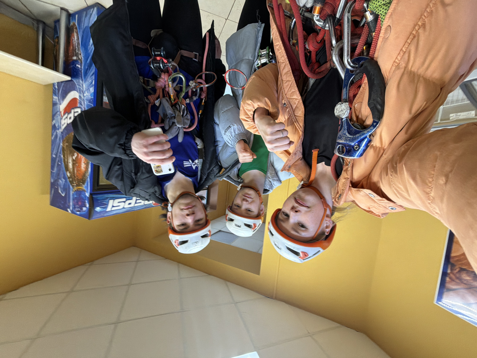 Three people, likely climbers, stand in an indoor space. The individuals wear climbing helmets and climbing harnesses with various carabiners and ropes attached. The person on the left holds a phone. The setting is possibly near a store or vending machine displaying Pepsi products. The wall color is yellow, and the floor is tiled. The specific location in Berezniki, Russia, is not explicitly evident from the image.