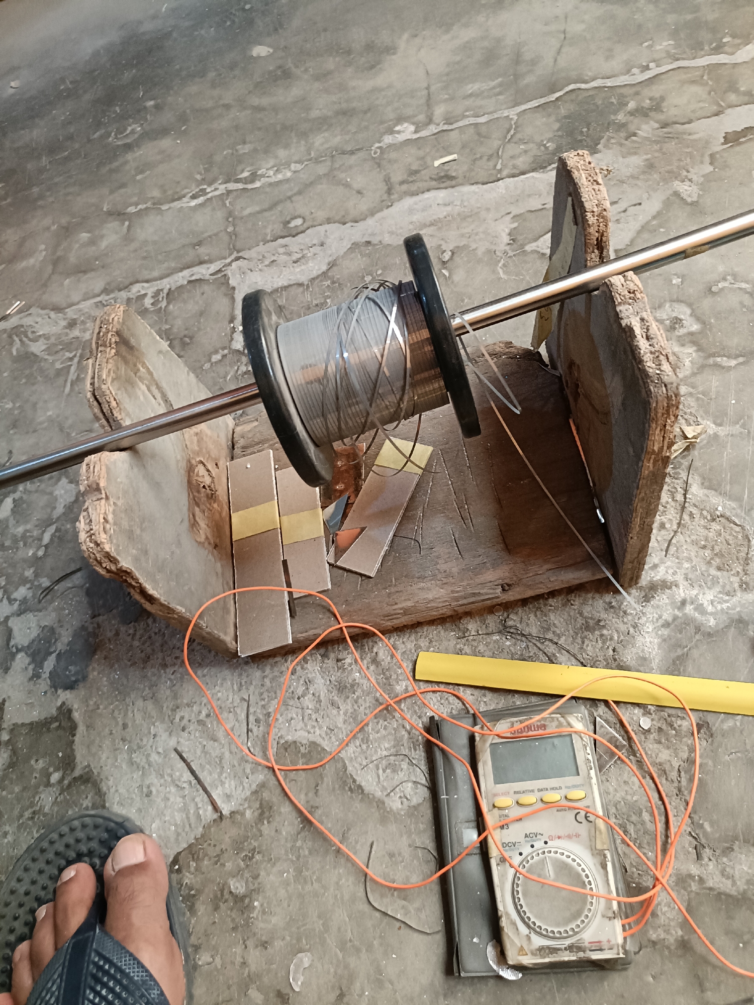 An outdoor, ground-level medium shot shows a collection of materials on a concrete surface. In the center is a wooden spool holder with a metal rod, holding a spool of thin wire. The spool is gray and appears to be made of plastic. A thin orange wire is loosely coiled around the spool holder and extends towards the lower right of the frame. To the right of the spool holder, a digital multimeter is visible, with its screen facing upward. The multimeter is white with yellow buttons and has the brand name "Sanwa" visible. An orange wire is also connected to the multimeter. A yellow plastic tube is positioned horizontally above the multimeter. In the lower left corner, a person's left foot wearing a dark sandal is visible. The environment appears to be outdoors, with a rough concrete ground. There are no specific landmarks or features to pinpoint the exact location within Telagamurni, Indonesia, but the scene suggests a practical or work-in-progress setting.