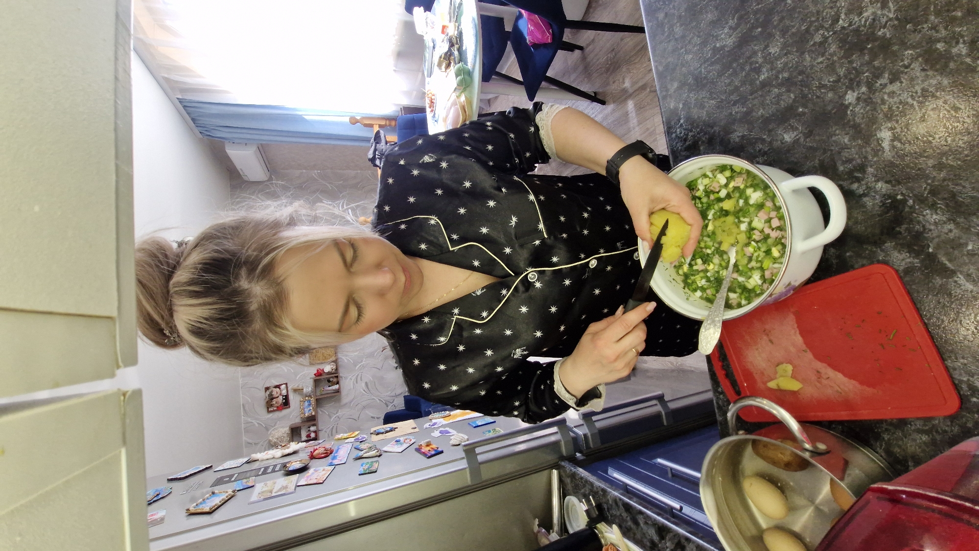 A woman with blonde hair tied in a bun is captured in a kitchen setting in Otchor, Russia, actively preparing food. She wears a black pajama-style top adorned with white star patterns and gold piping. Her expression is focused and calm as she looks down at her task, her eyes gently closed in concentration.
She is standing at a dark, speckled kitchen counter, adding ingredients to a large white pot. In her left hand, she holds a piece of a yellow vegetable, likely a cooked potato, and a small knife. The pot contains a mix of chopped green ingredients, possibly green onions, dill, and other vegetables, along with cubed ingredients that resemble ham or sausage and eggs, suggesting the preparation of a salad or cold soup like Okroshka. Next to the pot is a red cutting board with some scraps, and further to the right, a small metal pot containing whole eggs can be seen.
The background reveals a home environment. A refrigerator, covered with numerous colorful magnets, is visible to the left. Beyond the woman, a dining area with a table and blue chairs is partially visible, illuminated by natural light streaming through a window with light blue curtains. The walls feature a subtle patterned wallpaper. The bright light suggests it is daytime. No discernible text is visible on any objects in the image.