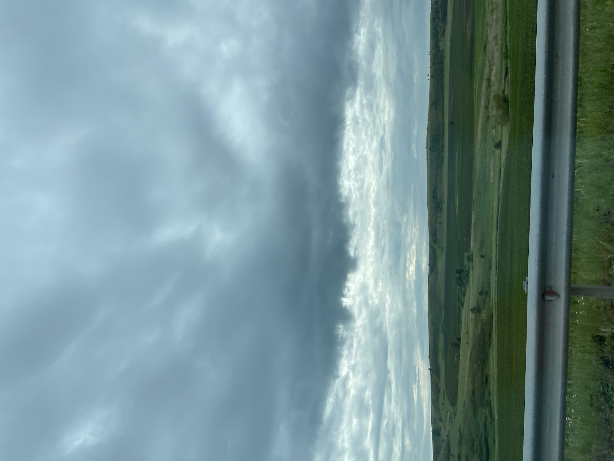 The image depicts a wide, open landscape under a heavily overcast sky. The terrain is characterized by rolling hills covered in various shades of green vegetation, suggesting agricultural fields or natural grasslands. In the foreground, a metal guardrail runs horizontally across the bottom of the frame, partially obscuring the view of the lower fields. The sky is filled with thick, dark clouds, casting a muted light over the entire scene. The overall impression is one of a somewhat somber and natural environment. The location is identified as Subkhankulovo, Russia, though no specific landmarks within the city are discernible in this general landscape shot. There are no people or discernible activity visible in the image.