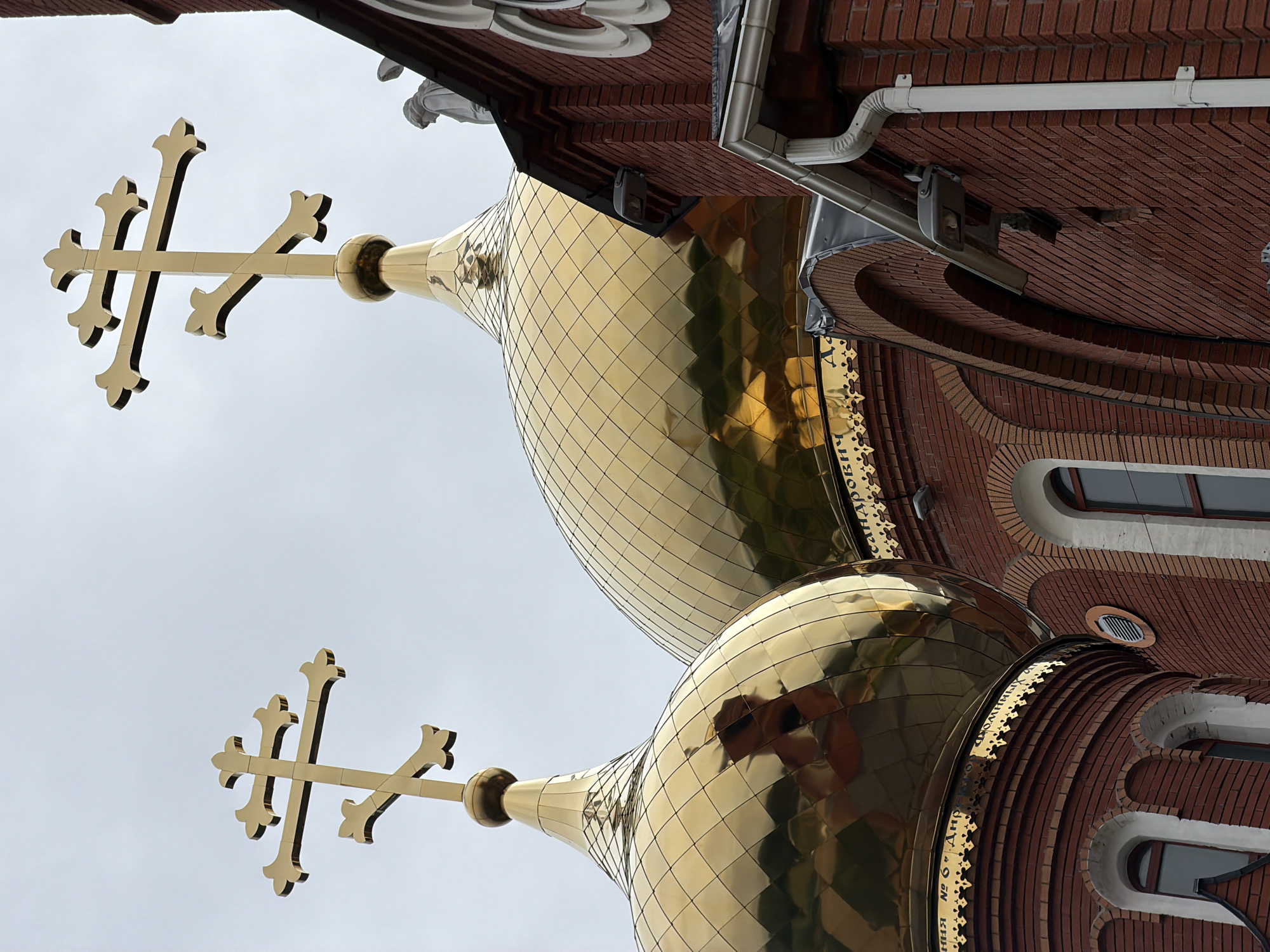 This is a low-angle, full shot of the golden domes and red brick facade of an Orthodox church, likely the Church of the Assumption of the Virgin Mary in Chaikovsky, Russia. Two ornate golden crosses, characteristic of Eastern Orthodox churches, top the conical domes. The domes are segmented with a diamond pattern, reflecting the overcast sky and surrounding elements. The church's red brickwork is visible below, with arched windows and decorative white trim. A white gutter system runs along the edge of the roof. In the background, a statue of a person, likely a saint, is partially visible near the roofline on the right side of the image. The overall scene is one of religious architecture against a muted sky.