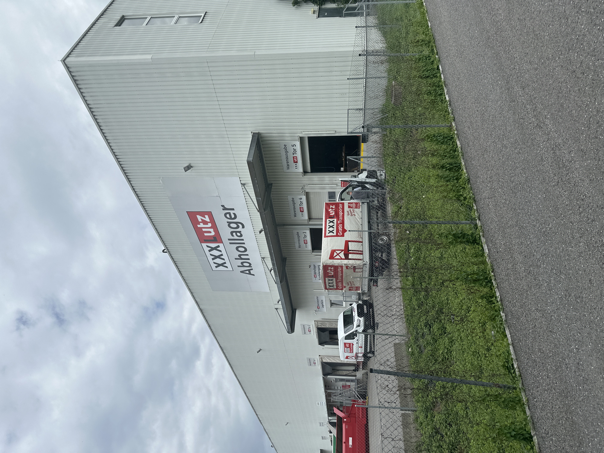 This is a wide, eye-level, outdoor shot of a large, modern industrial building with a light-colored, corrugated metal facade. A large white sign with red lettering is prominently displayed high on the building, reading "XXX Lutz Abholager" which translates to "XXX Lutz Collection Point." Below this main sign, there are several loading docks with gates and signage indicating "Warenausgabe" (Goods Issue) and gate numbers such as "Tor 3" and "Tor 4."
In front of the building, a chain-link fence runs horizontally across the foreground, with overgrown green vegetation behind it. Several vehicles are parked or moving in front of the loading docks. A white van with "XXX Lutz Gratis Transporter" branding is visible, as is a larger, red truck with similar branding and a prominent graphic of a red chair. Two individuals, appearing to be employees, are interacting near the white van. One is standing by the open driver's side door, and the other is near the back of the van. A red industrial dumpster or container is also present near one of the loading docks. The sky is overcast with visible clouds, suggesting a cloudy day. The location is the XXX Lutz logistics center in Lauterach, Austria.