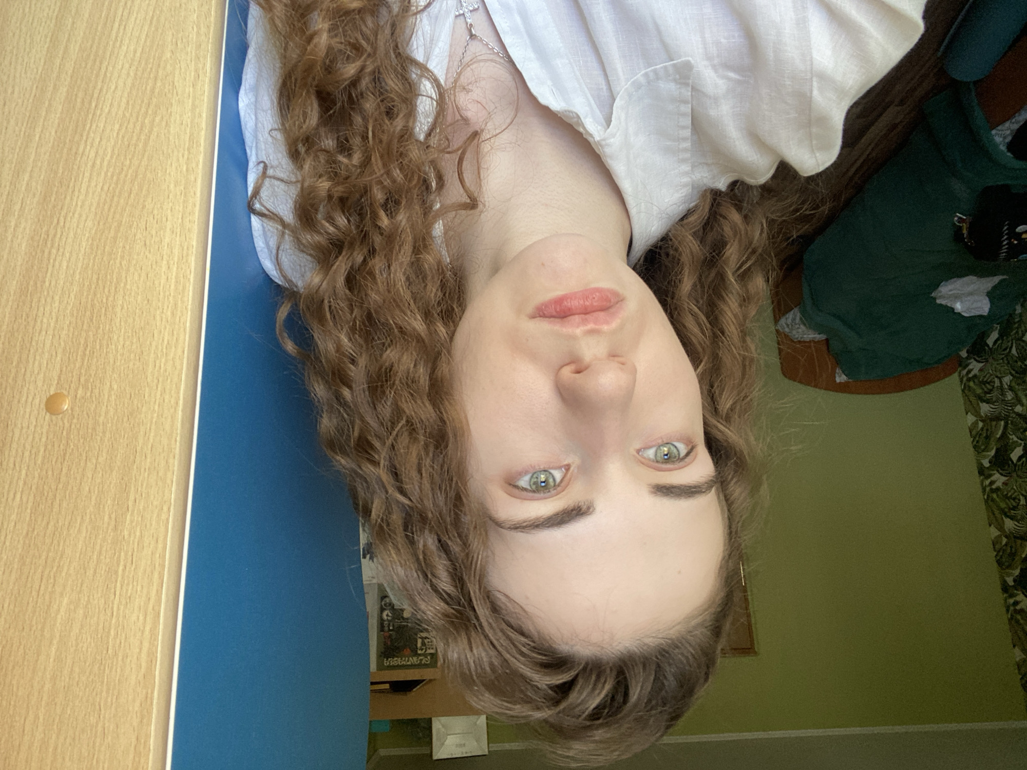 The media file is a portrait of a young person with curly brown hair, wearing a white shirt, and a silver cross necklace. Their face is tilted upwards, and they have striking green eyes and light skin. The image is oriented such that the person's head is at the bottom.
The setting appears to be indoors, with a wooden surface on the left, a blue surface in the middle, and a green wall on the right. There are also hints of a darker fabric and a patterned surface in the background, possibly indicating furniture or other room decor. The lighting suggests it is daytime, but no specific time can be determined.
The activity appears to be a selfie, with the camera angled upwards towards the subject's face. The subject's expression is neutral.
Visible text includes "PLANETA" which appears on a piece of media in the background.
The location context is Zavodoukovsk, Russia.