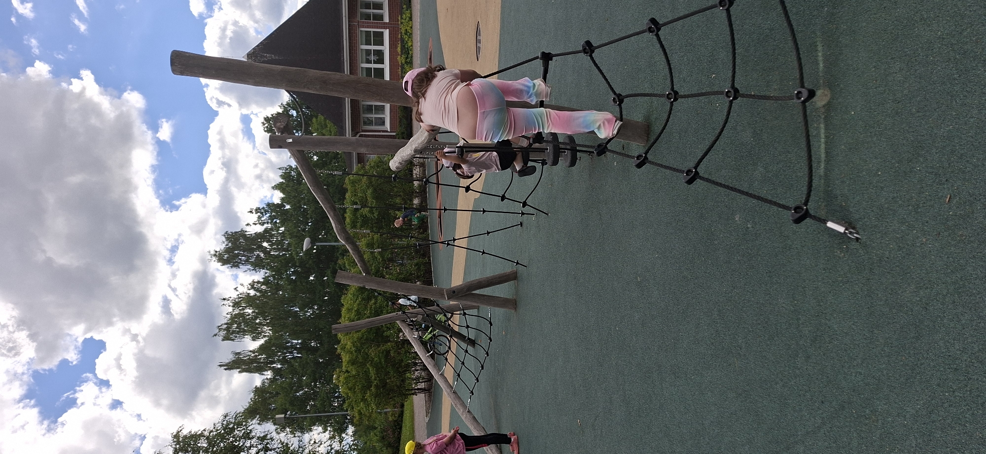 The image depicts a playground area in Lahti, Finland. The playground features a large wooden climbing structure with ropes and nets. Several children are present, engaged in activities on the equipment. In the foreground, a child wearing a pink shirt and rainbow-colored pants is climbing a rope net. To the left, another child in a pink shirt and yellow helmet stands on the green rubberized ground. In the background, there is a brick building with windows, surrounded by trees and green bushes. The sky is partly cloudy with white cumulus clouds against a blue sky. A bicycle is visible leaning against the wooden structure. The ground surface is a textured green, with beige and light brown colored areas demarcated by lines.