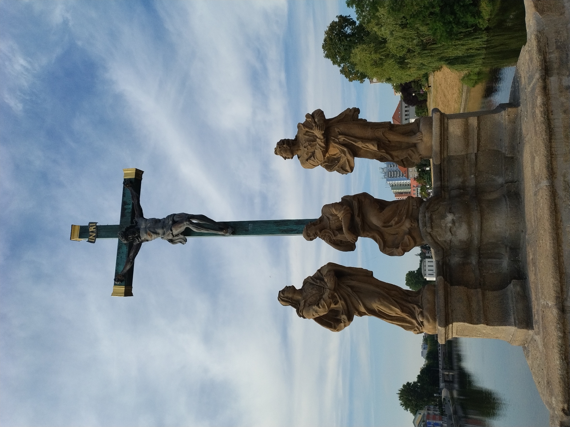 The image shows a statue of Jesus Christ on the cross, with three accompanying statues of women at the base. The statue is located on the Písek Stone Bridge (Kamenný most) in Písek, Czechia. The bridge spans the Otava River. The sky above is blue with scattered white clouds. The surrounding environment includes trees, buildings, and the river. The statue of Jesus is dark in color, with the inscription "INRI" on the cross. The three women are depicted in flowing robes, with serene expressions. The stone bridge and its statues appear weathered, suggesting age. The overall scene conveys a sense of religious solemnity and historical significance.