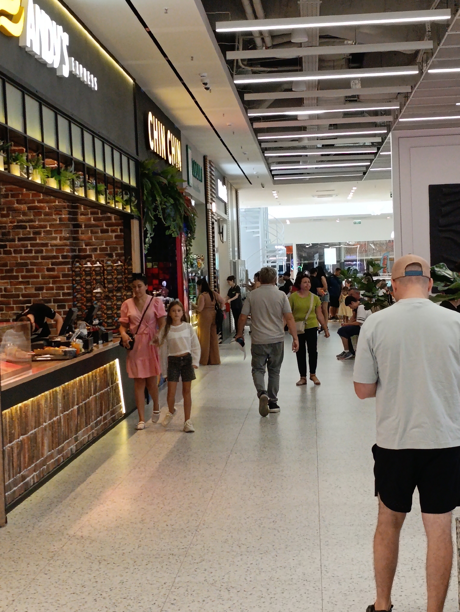 The image shows an indoor marketplace or shopping mall with a bright, modern aesthetic. In the foreground, a person wearing a light blue t-shirt and black shorts is walking away from the camera, their back mostly to the viewer. To the left, a brick wall with shelves displays items, and a counter is visible with food and drinks. Three people are walking past the counter: a woman in a pink dress, a young girl in a white top and shorts, and a man in a grey t-shirt and jeans. Further into the mall, other shoppers are seen walking and standing. The ceiling features a grid of fluorescent lighting. The overall scene suggests a commercial area with multiple shops and foot traffic. The provided context suggests this location is in Chișinău, Moldova.