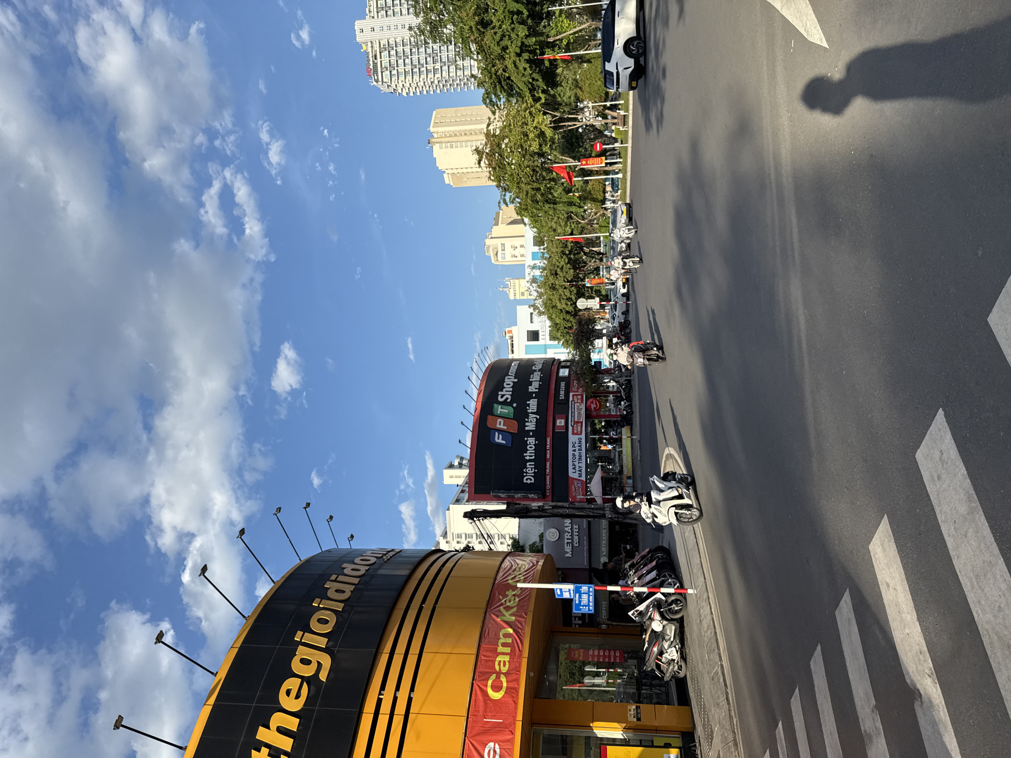 In Nha Trang, Vietnam, a street scene unfolds with a clear blue sky dotted with white clouds. On the left, a large, curved yellow and black building displays signage that reads "thegioididong.com" and "Cam Ket". To its right, a large billboard for "FPT Shop" with the slogan "Điện thoại - Máy tính - Phụ kiện - Gia" is visible. Various shops and businesses, including "METRAN COFFEE", line the street. A row of parked motorcycles is present on the sidewalk, with one white motorcycle rider on the road in the foreground. Other motorcycles and cars are navigating the intersection. Tall residential buildings and lush green trees form the backdrop. A crosswalk is marked on the asphalt road. The shadow of a person standing on the sidewalk is cast across the road.