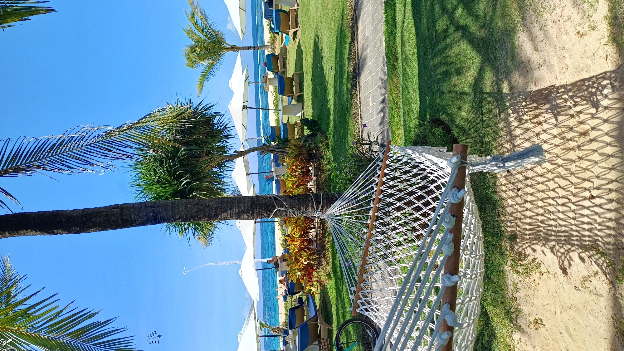 The scene shows a hammock stretched between a palm tree and a wooden post, indicating a relaxed or recreational environment. The location features a beach with sun umbrellas and chairs, suggesting a tourist area in Kuta Selatan, Indonesia. The sky is clear and blue, indicating good weather. There are plants and greenery surrounding the hammock, providing a natural and aesthetically pleasing environment.