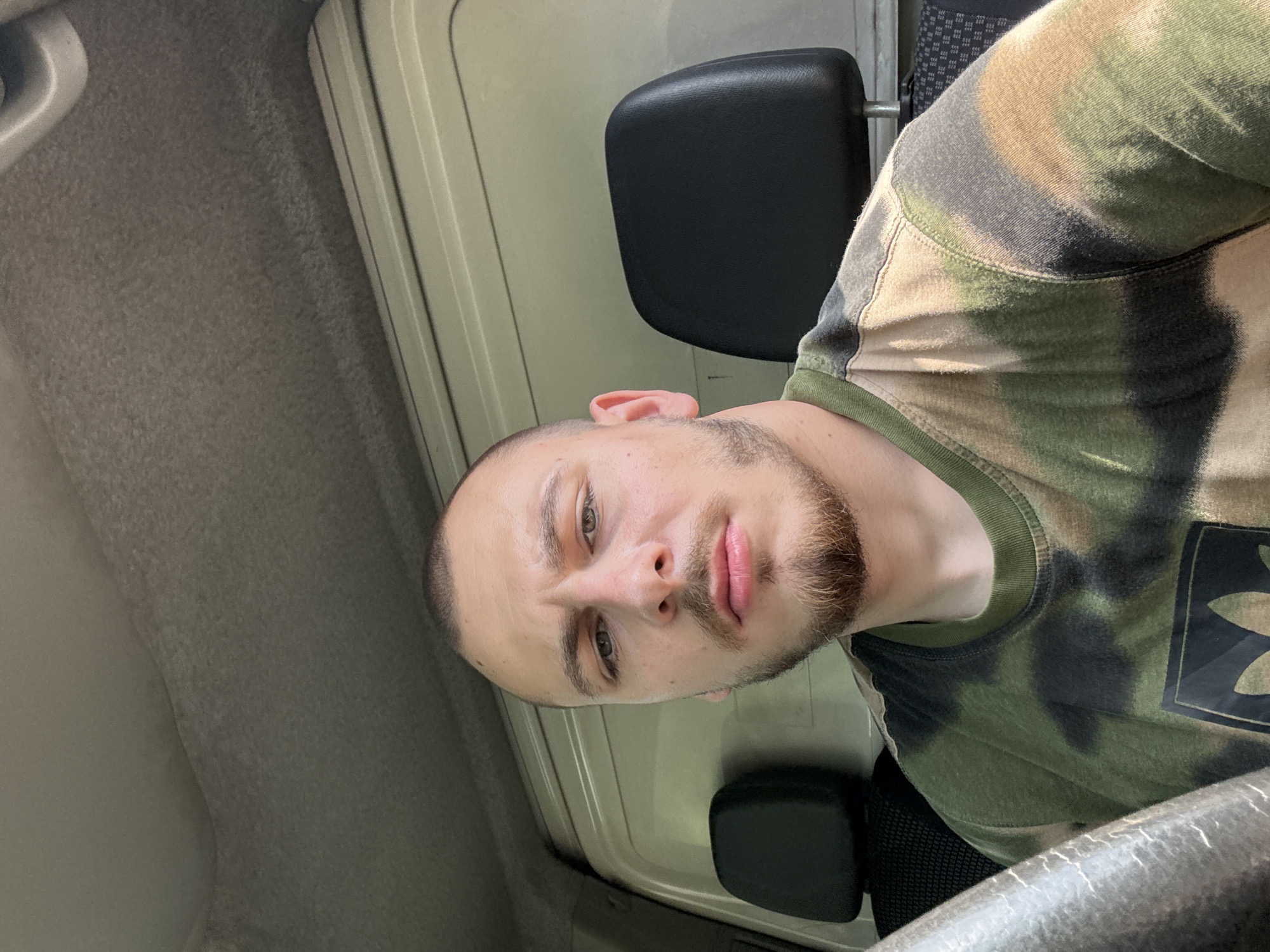 A man is captured in a close-up, rotated portrait from inside a vehicle, likely the cab of a truck or van, in Stara Zagora, Bulgaria. He has a light complexion, short dark hair, and a neatly trimmed light brown beard and mustache. His brown eyes are directed towards the camera, and his lips are slightly parted, conveying a neutral or slightly pensive expression.
He is wearing a camouflage-patterned t-shirt in shades of green, brown, and black, featuring a dark, stylized logo on the chest that is difficult to discern. The interior of the vehicle features utilitarian design with muted grey and beige headliner and wall panels. A black headrest is visible behind him, and the edge of what appears to be a steering wheel or dashboard is in the foreground. The lighting is soft and even, suggesting an indoor setting or diffused natural light during the daytime.