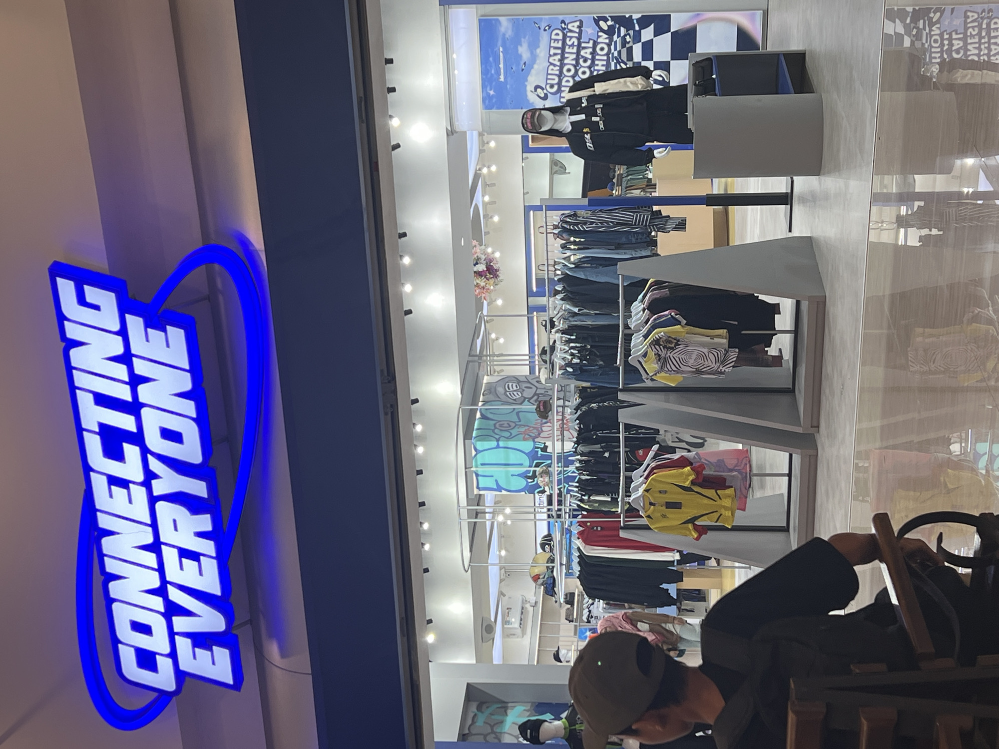 This image depicts the exterior of a clothing store named "CONNECTING EVERYONE" located in Bandung City, Indonesia. The store features a prominent blue neon sign with white lettering. Inside the store, racks of clothing are visible, including t-shirts and jackets. A mannequin wearing a black jacket and a cap stands near the entrance. The flooring appears to be reflective. In the foreground, a person wearing a black shirt and a dark cap is seated on a chair, facing away from the camera.