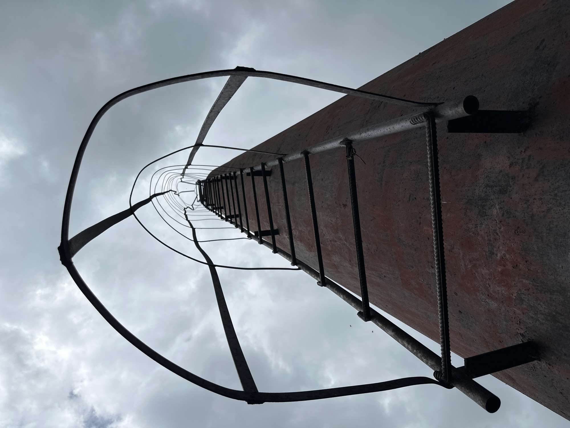 An upward perspective shot of a cylindrical structure, likely a smokestack or silo, with a metal ladder ascending its exterior. The ladder features a protective cage made of curved metal bars that extend upwards and enclose the ascending path. The structure itself appears weathered, with a dark reddish-brown hue and signs of rust and wear. The sky above is overcast with clouds. No people are visible in the frame. The image is taken in an outdoor environment, with no specific urban landmarks identifying the location beyond "Unknown, Unknown".
