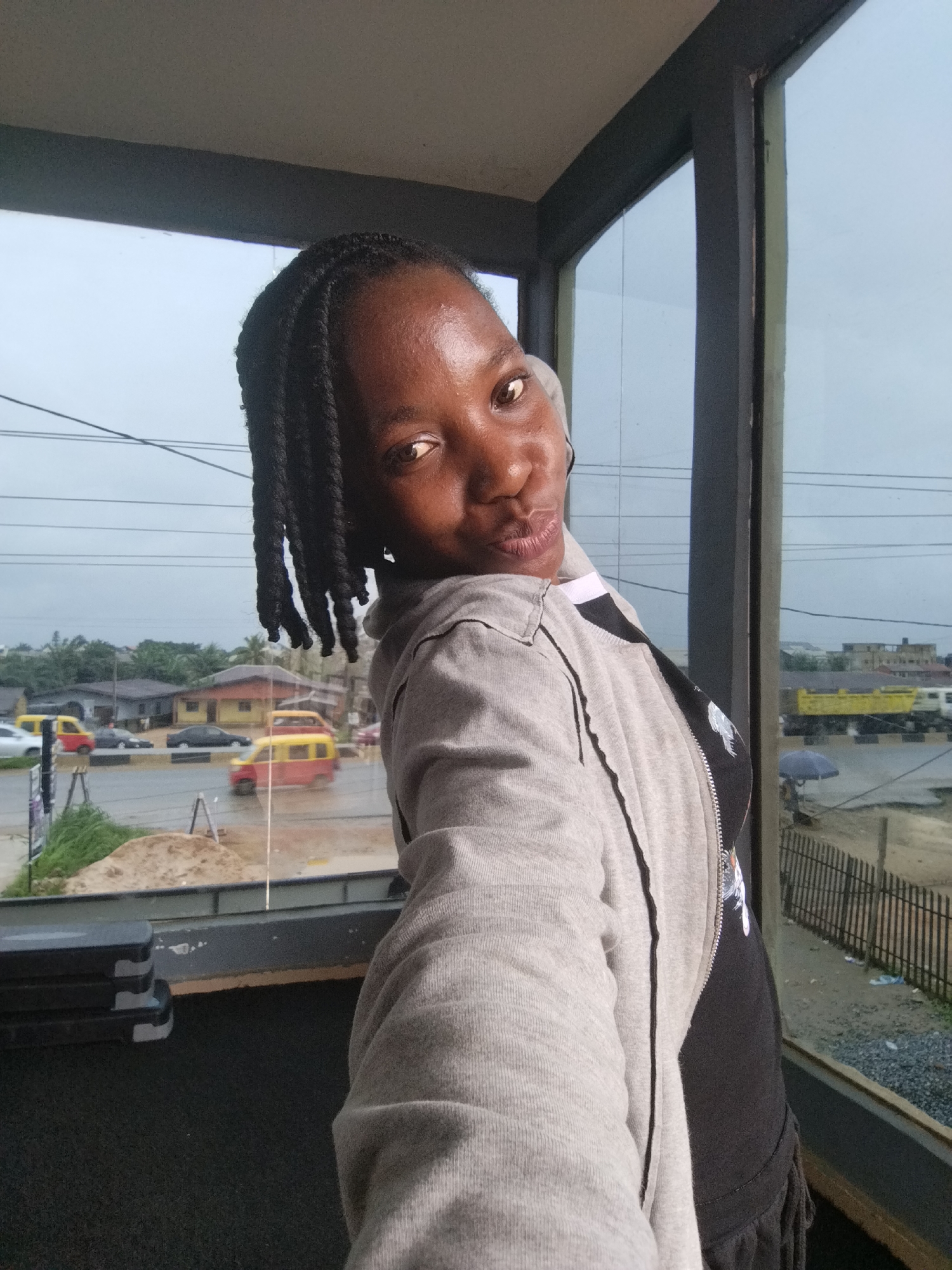 A woman with braided hair is taking a selfie in front of a large window. She is wearing a grey zip-up hoodie over a black t-shirt. The hoodie is open, revealing the t-shirt which has a graphic print on the chest. She is looking at the camera and puckering her lips. The window overlooks a street with several vehicles, including yellow and red mini-buses, cars, and a yellow truck. Buildings and trees are visible in the background, suggesting a semi-urban or rural setting. The sky appears overcast. The area below the window seems to be an indoor space, possibly a gym, with a black floor and stacked exercise step platforms on the left side. The scene suggests a casual moment captured from within a building. The context provided indicates the location is Uselu, Nigeria.
