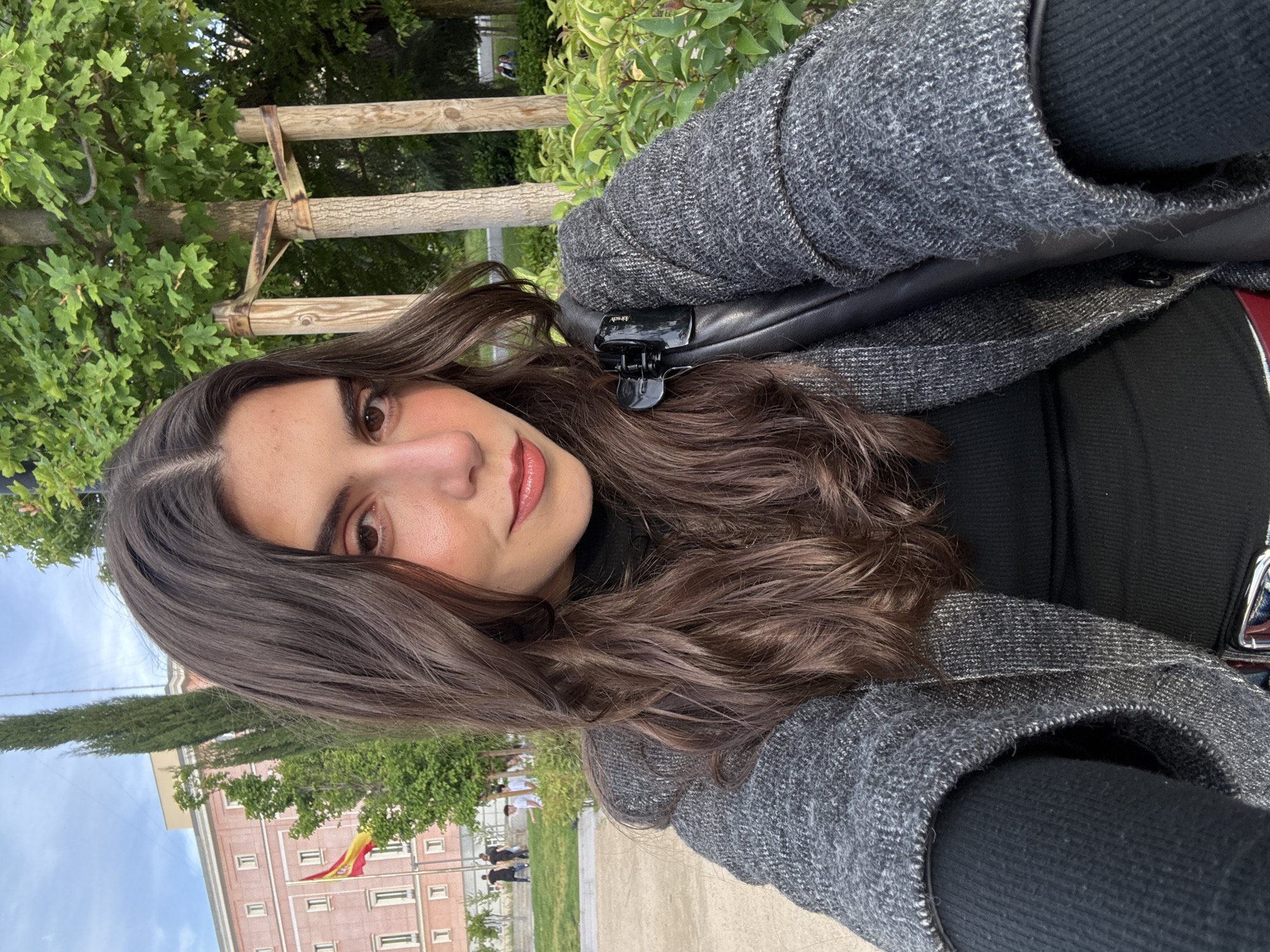 A medium close-up portrait of a woman with long brown hair, wearing a dark top and a gray tweed jacket, is rotated counter-clockwise. Her hair is partially held back with a black hair clip. The background includes a building flying the Spanish flag, trees, and a wooden fence. The setting appears to be a park or plaza in Madrid, Spain. A few blurred figures can be seen in the distance. The ground is paved.