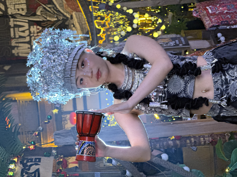 An East Asian female subject is centrally positioned, facing slightly right, within a vibrantly lit, decorated outdoor or semi-outdoor setting in Yuzhong District, China. She wears an elaborate silver-colored headpiece composed of layered metallic elements, reminiscent of traditional ethnic headdresses. Her hair is styled in two thick, black braided pigtails, each adorned with silver-colored accents. The subject's attire includes multiple layered silver-colored necklaces, long dangling earrings, and a black crop top featuring white circular patterns, exposing her midriff which displays a silver-colored navel piercing. A matching patterned skirt is partially visible below. She holds a small, red, djembe-style hand drum with black, blue, and white decorative patterns in her raised left hand. Her right hand is also raised to chest level, palm forward, in a gestural pose. Her face exhibits glitter on the cheeks and pink lip gloss, with a neutral expression. The background is dimly lit but features numerous warm yellow string lights, star-shaped decorations, and a red lantern. To the left, a section of a palm tree is visible, wrapped with colorful string lights, alongside a white banner displaying the red Chinese characters for '宾馆' (hotel/guesthouse). A colorful, patterned textile hangs vertically on the far right.