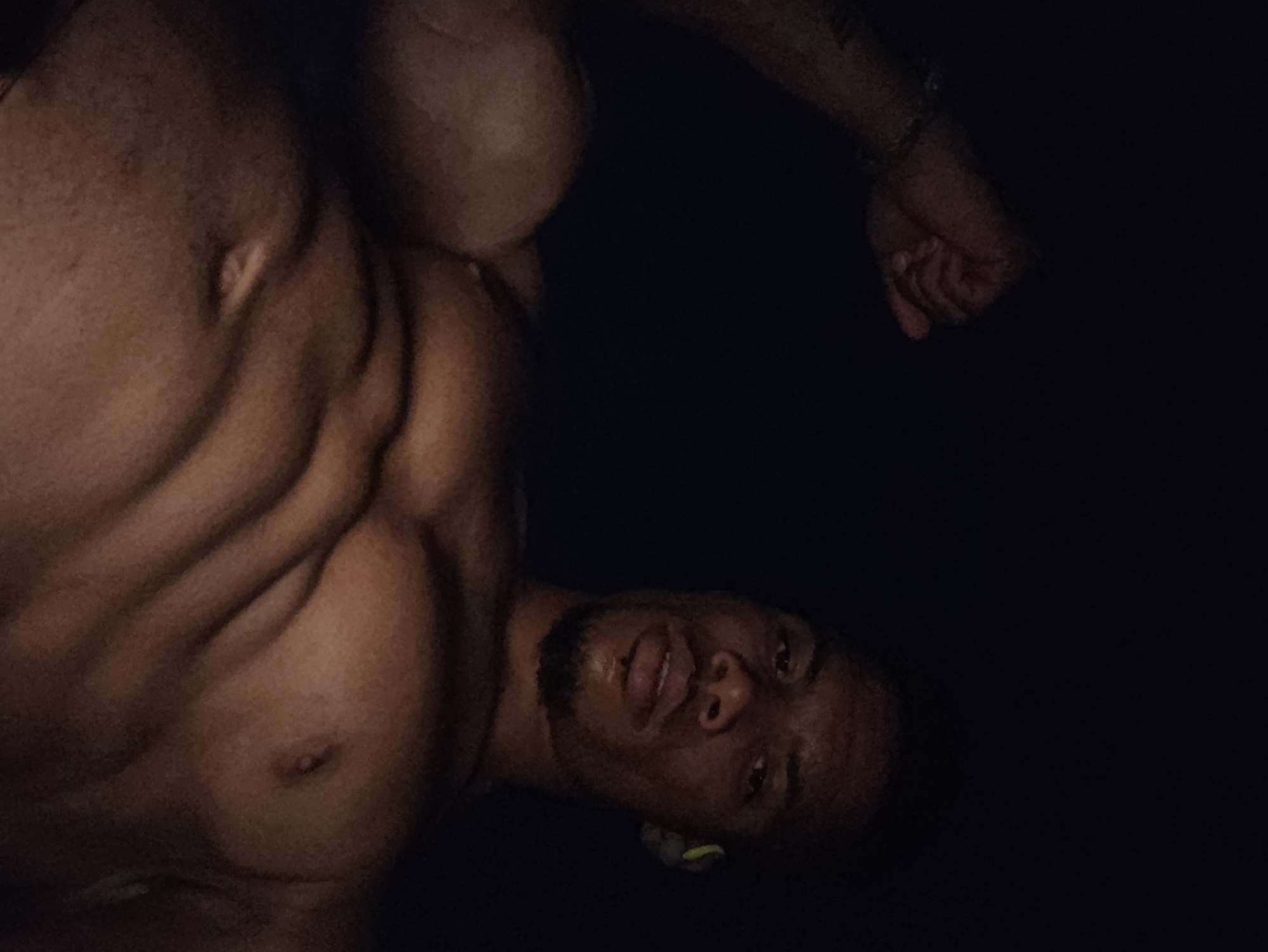 The image is a low-angle, close-up, self-taken photograph of a muscular, shirtless Black man with a goatee and a small beard. He is wearing wireless headphones in his right ear. His left arm is flexed and raised, with his fist clenched. The man is looking directly at the camera with a neutral expression. The background is dark and indistinct, with no discernible urban or natural features. There is no information in the image or provided context to suggest that this image is located in Varna, Bulgaria.