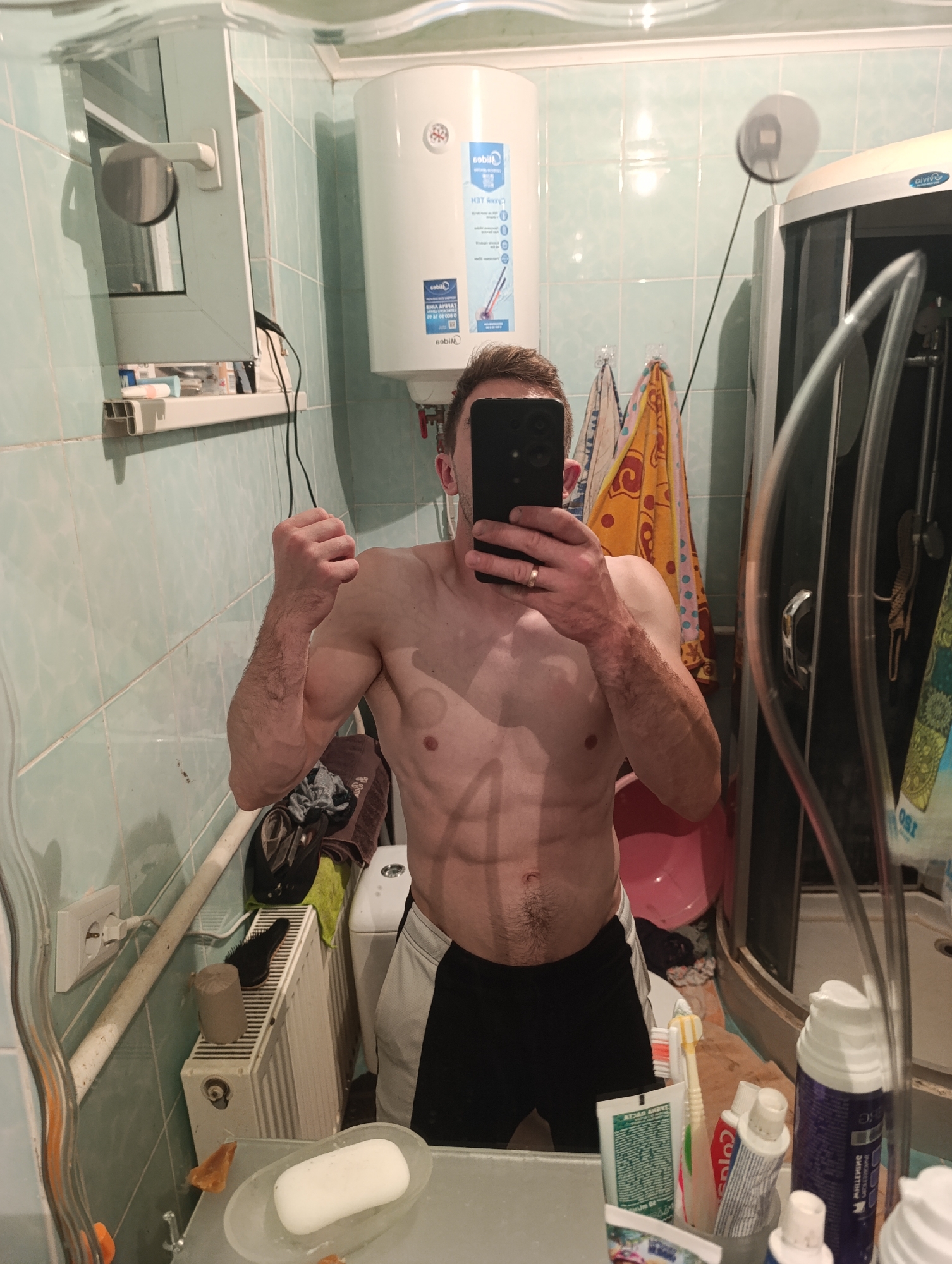 In a bathroom, a man is taking a selfie in a mirror. The bathroom has light blue tiled walls and a white ceiling. There is a large white water heater mounted on the wall, with a blue and white label on it. To the right, a corner shower stall with a transparent door is visible. The man is shirtless, wearing black and white athletic shorts, and is flexing his arm. His abs are clearly defined. In the mirror, a shelf is visible on the left, holding various toiletries. Below the shelf, a white radiator is present, with a black hairbrush and a roll of toilet paper on it. A white electrical outlet is also on the wall. The mirror shows a reflection of the man, the bathroom fixtures, and items on the sink counter, including a bar of soap in a clear dish, toothbrushes, and toothpaste.