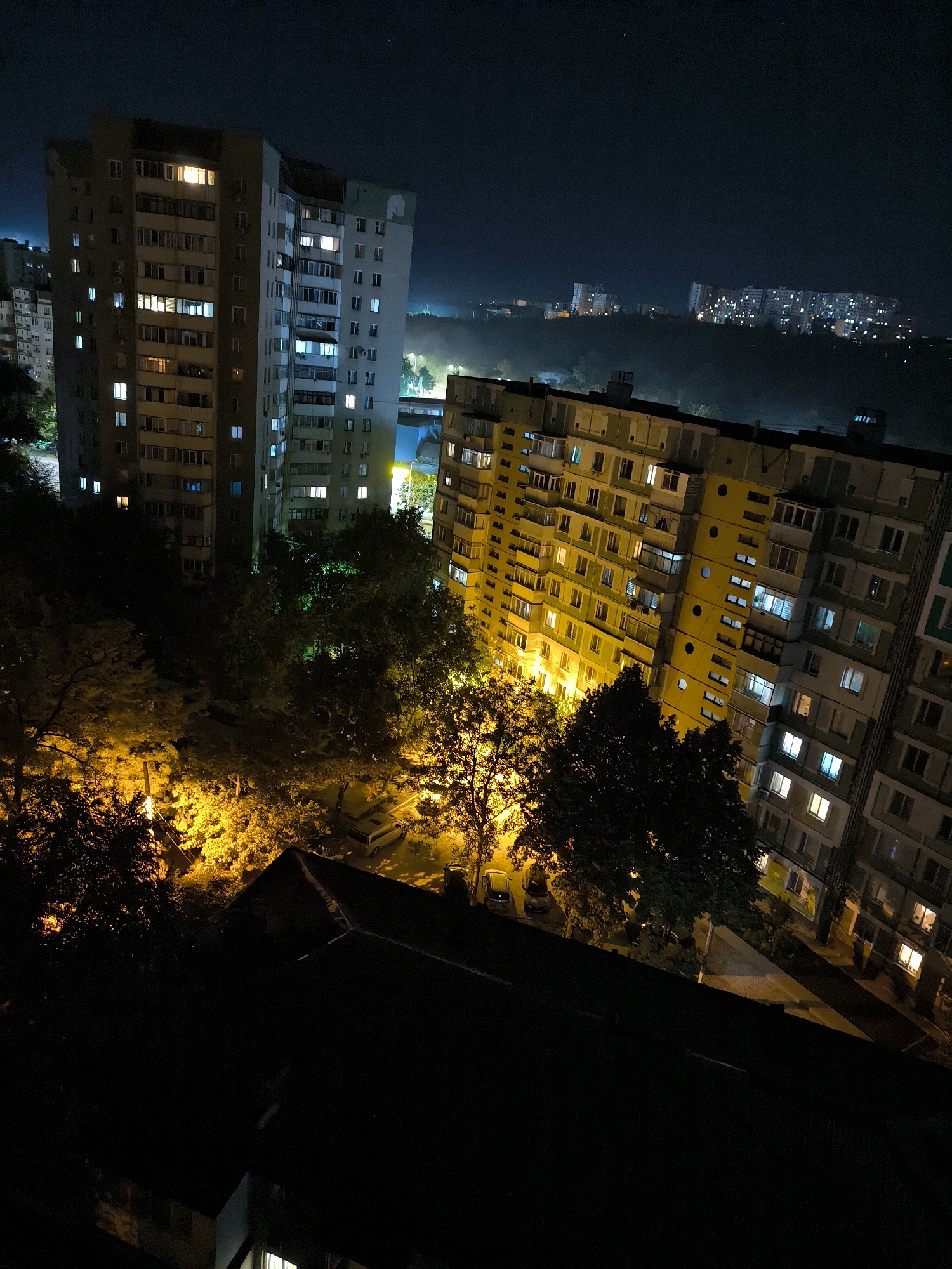 This is a nighttime aerial shot overlooking a residential area in Chișinău, Moldova. The foreground is dominated by the dark silhouette of a rooftop, obscuring the view of lower structures. Beyond this, several apartment buildings are visible. To the left, a tall, grey building with numerous lit windows stands out against the dark sky. In the center and right, a longer, yellowish apartment complex with many illuminated windows stretches across the frame, appearing slightly tilted. Below the buildings, trees are visible, some illuminated by warm, artificial light, casting a yellow glow. In the distance, more apartment buildings are faintly visible, their lights twinkling against the dark, hilly landscape. The overall scene depicts a typical urban environment at night, with the distinct architecture of Soviet-era apartment blocks characteristic of Chișinău.