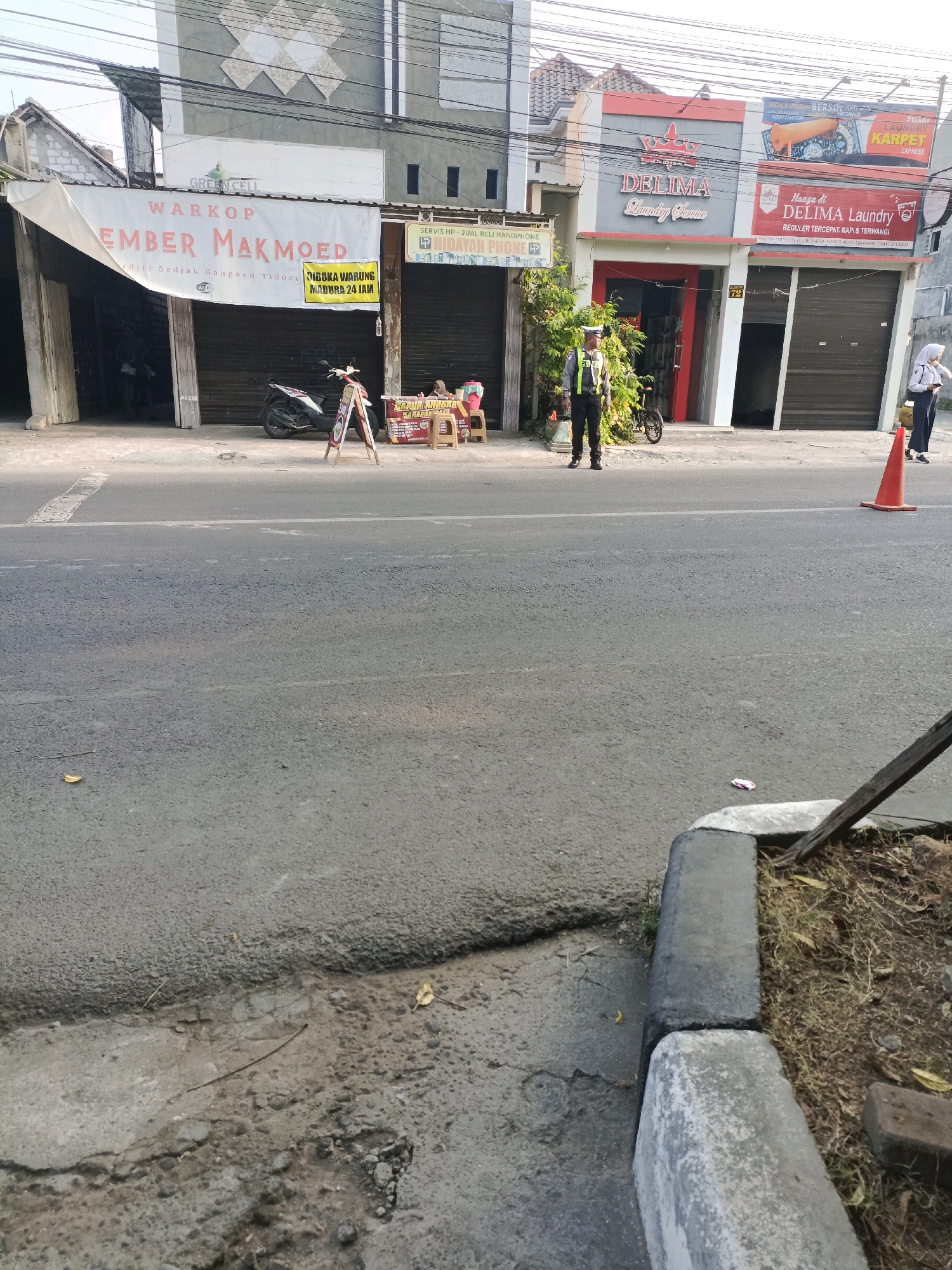 This image depicts a street scene in Sukomulyo, Indonesia, focusing on the facade of several businesses. In the foreground, a paved road is visible with a cracked concrete curb and sparse vegetation on the right.
On the left side of the street, a warung, identified by a banner reading "Warkop Ember Makmoed," is partially visible with its shutters down. A motorcycle is parked in front of it. To the right of the warung, a smaller sign in yellow indicates "Dibuka Warung Madura 24 Jam," suggesting a food or drink establishment operating around the clock.
Further to the right, a building with red and gray accents houses "DELIMA Laundry Service." A uniformed police officer stands on the sidewalk in front of this establishment, facing towards the street. Next to the laundry service, another sign advertises "PUSAT LAUNCHER KARPET EXPRESS."
On the far right of the scene, a woman in a white headscarf and dark clothing is walking away from the camera. An orange traffic cone is placed on the road near her. The buildings appear to be low-rise commercial structures, typical of a local market or street shopping area. The overall atmosphere suggests a moderately busy daytime scene in a neighborhood commercial district.