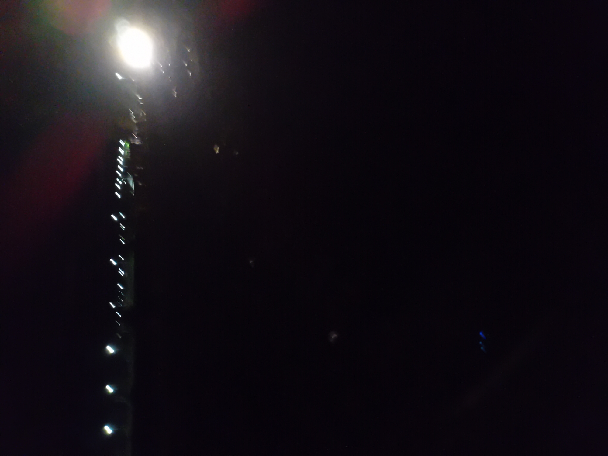 This image depicts an outdoor scene at night, likely in Jauro Yinu, Nigeria, characterized by darkness and scattered artificial lights. The most prominent feature is a bright, out-of-focus light source on the right side of the frame, possibly a headlight or a powerful lamp. A line of blurred white lights stretches across the upper portion of the image, suggesting streetlights or lights from buildings in the distance. The ground appears to be a dark, flat surface, with some scattered, faint glints of light, potentially from reflective debris. The overall impression is one of a dimly lit, open area with a strong artificial light source dominating the right side.